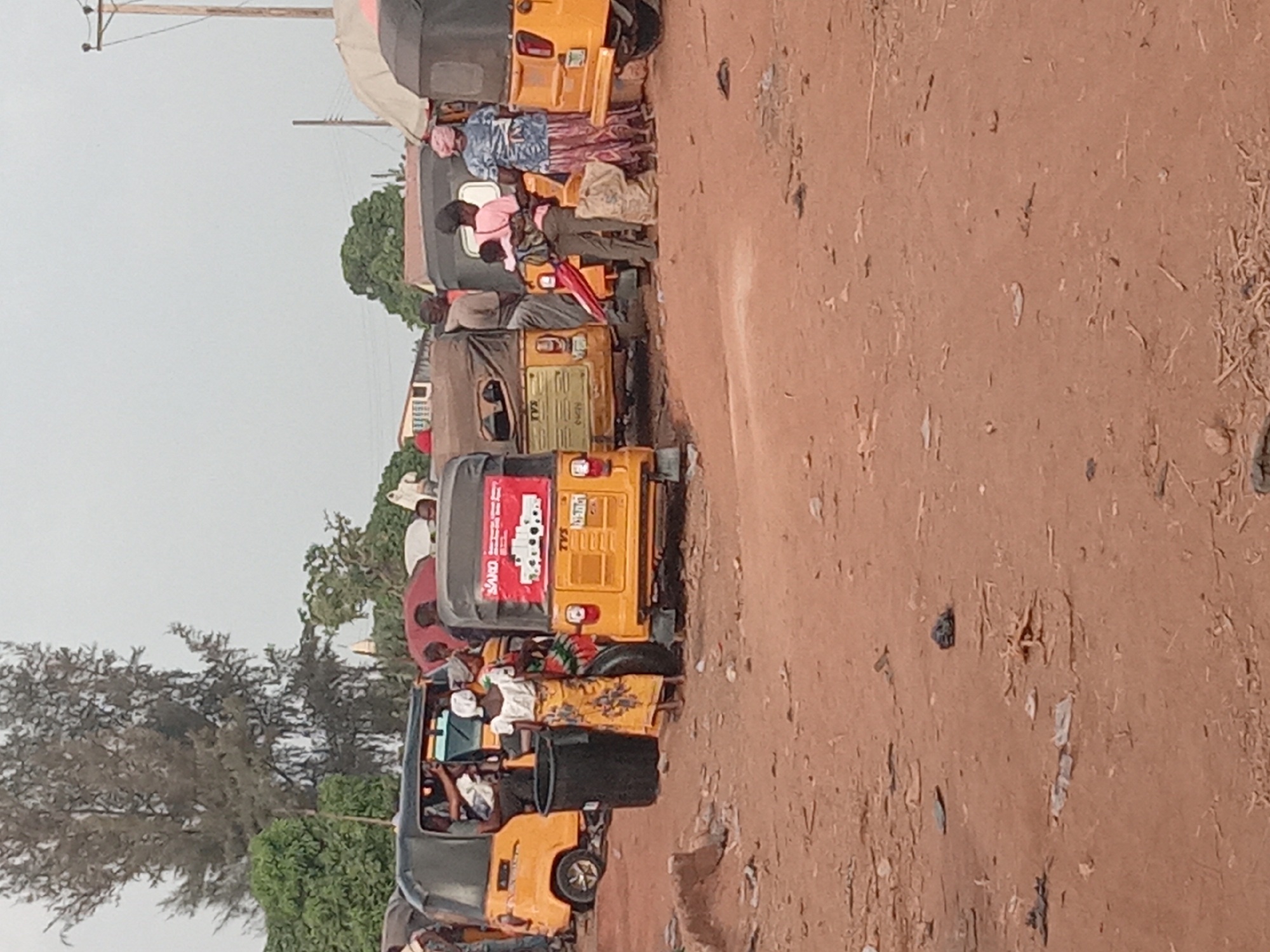 The image depicts a row of auto rickshaws, colloquially known as "keke," lining a dirt road in Kakuri Gwari, Nigeria. Several passengers are sitting in the vehicles. The environment includes trees and foliage. A utility pole with electrical lines is visible overhead. Litter and debris are scattered on the ground along the edge of the road. One of the auto rickshaws has an advertisement.