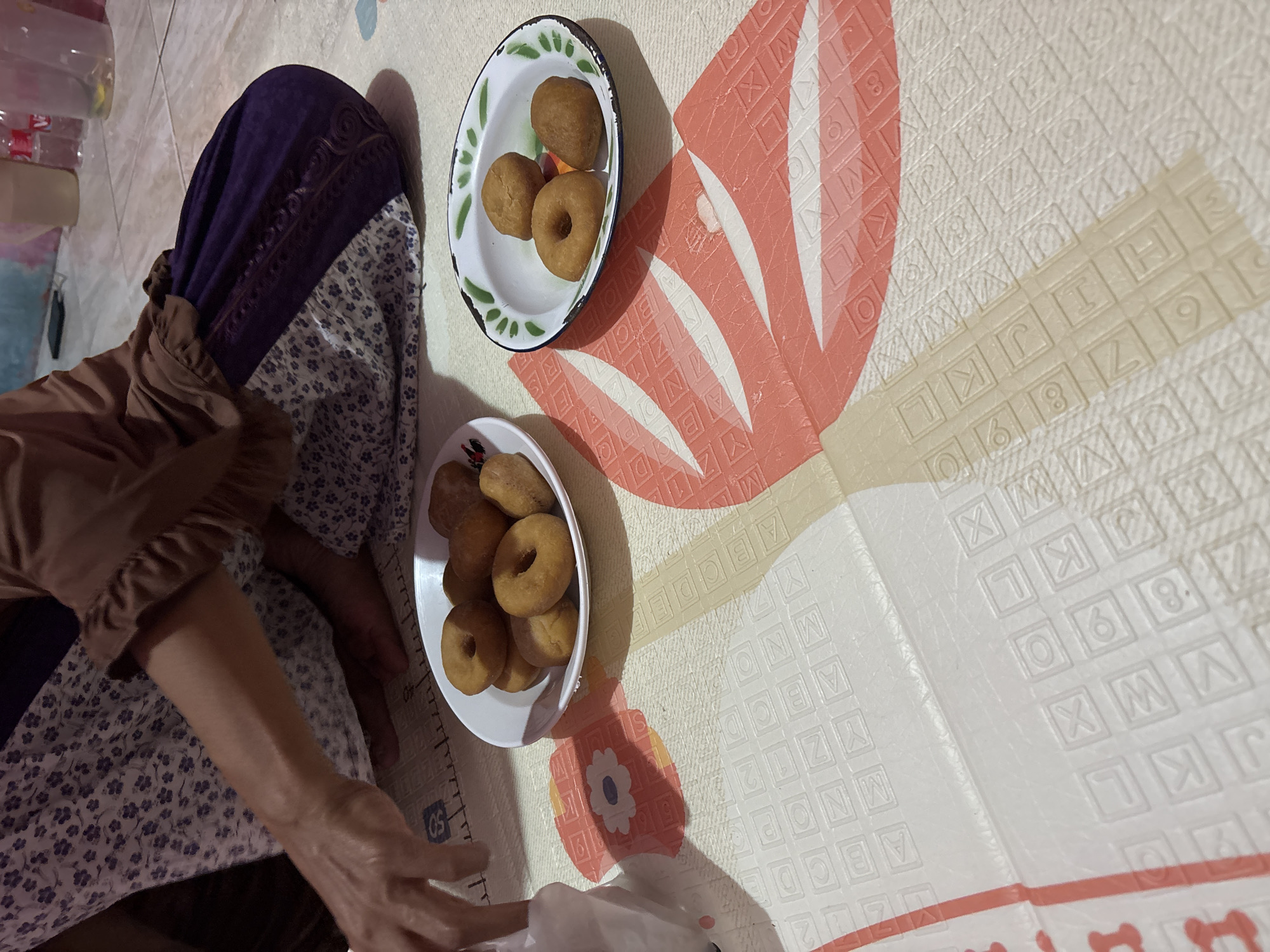 The image depicts a close-up, low-angle view of a person sitting on a mat, with two plates of donuts in front of them. The person, only partially visible from the elbow down, is wearing a brown long-sleeved shirt and a patterned purple and white garment. Their left hand is reaching towards a white plastic bag.
The first plate, positioned to the left, is white and contains approximately seven golden-brown donuts, some ring-shaped and some solid. The second plate, to the right, is a vintage-style metal bowl with a green rim, containing three similar golden-brown donuts.
The mat the person is sitting on appears to be a playmat with an alphabet and numerical pattern, interspersed with large pink and orange abstract floral designs. The background is blurred, showing parts of a wall and some bottles. The lighting suggests an indoor setting, possibly a home. The image does not provide any specific information about the location within Telagamurni, Indonesia, beyond it being an indoor domestic environment.