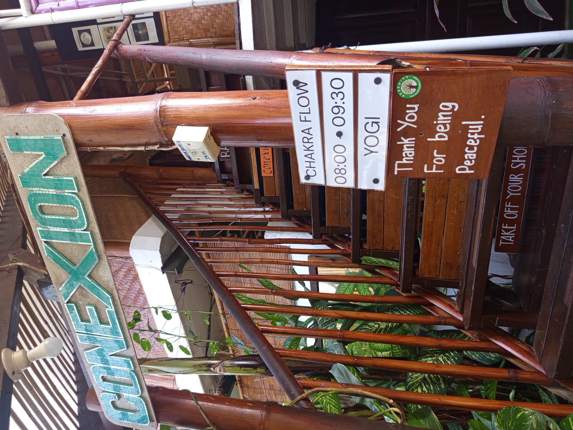 The image depicts a view of Conexión, a venue in North Kuta, Indonesia, specifically focusing on a staircase. The staircase is constructed from dark wood and bamboo, with green plants growing along the side. Several signs are attached to the wooden structure. One sign displays the words "CHAKRA FLOW" with the time "08:00 - 09:30" and "YOGI." Another sign expresses "Thank You For being Peaceful," including the logo for "Serenity Eco Guesthouse & Yoga." Additionally, a sign says "TAKE OFF YOUR SHO" and a partial view of another sign that has "Conexi."