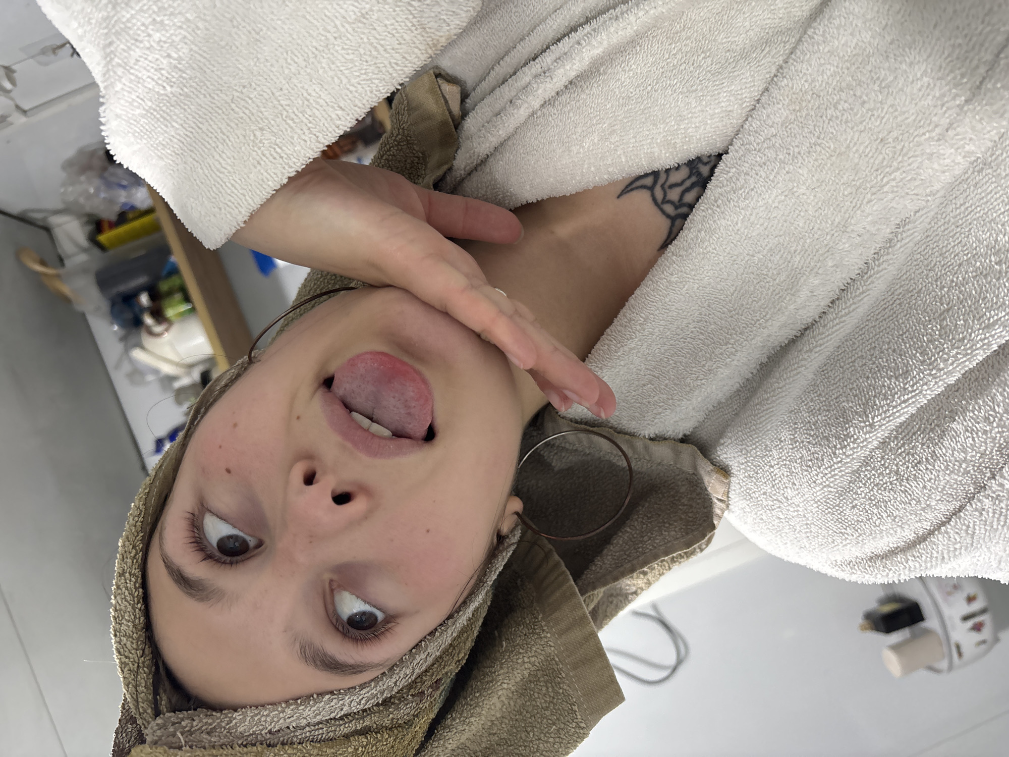 A woman is posing for a selfie in a bathroom. She is wearing a white robe and a brown towel around her head. She has her tongue sticking out and is holding her hand to her throat. A tattoo is visible on her chest. There is a sink and some items on the counter behind her. It is not possible to determine the location of the city of Фоки, Russia.