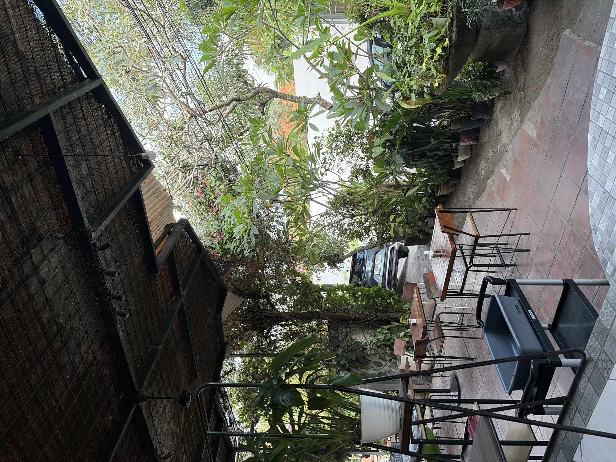 An outdoor seating area of a cafe in North Kuta, Indonesia is depicted, characterized by lush greenery and a rustic, covered roof structure. The roof is made of woven material stretched over metal beams, with some lighting fixtures visible. Several tables and chairs, mostly made of wood and metal, are arranged on a tiled floor. A large potted plant sits on a shelf to the left, and a serving cart with a gray bin is positioned in the foreground. Beyond the seating area, a variety of potted plants line the path. In the background, a dark-colored car is parked, and a white wall is visible. The overall atmosphere is natural and relaxed, suggesting a pleasant dining or resting spot.