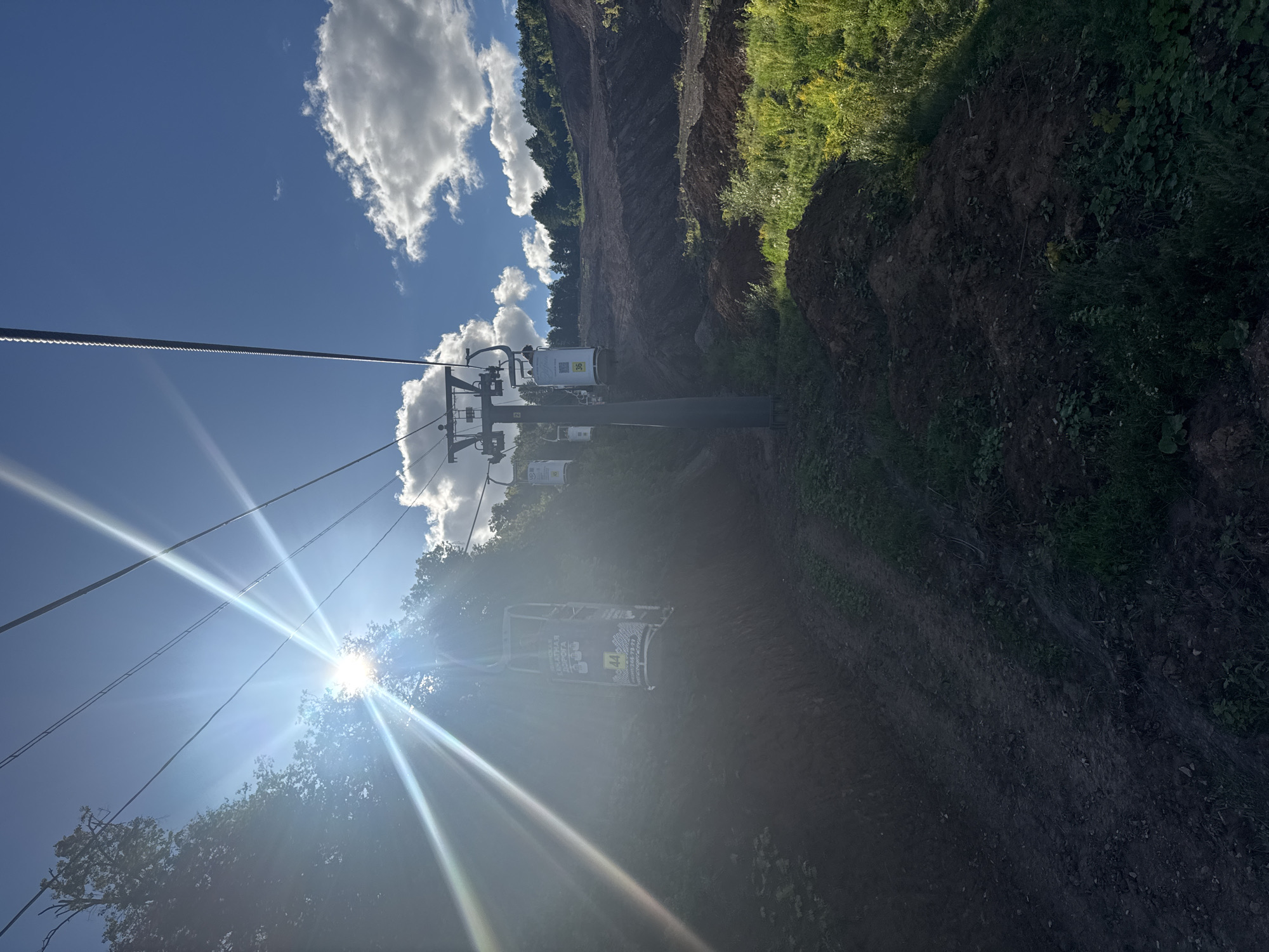 The image shows a ski lift with several cabins against a bright blue sky with scattered white clouds. The sun is shining intensely, creating lens flare and streaks of light across the upper left portion of the frame. The ski lift poles and cables are visible, stretching upwards and across the image. Some cabins are numbered, such as "44" and "36". The ski lift is situated on a wooded hillside, with trees visible in the background. The foreground and midground depict a rugged, uneven terrain with exposed soil and patches of grass and vegetation, suggesting a construction or landscaping area. The overall scene appears to be outdoors during the daytime, with the sun indicating it is likely midday or early afternoon. The location is not specified in the image or provided crops.