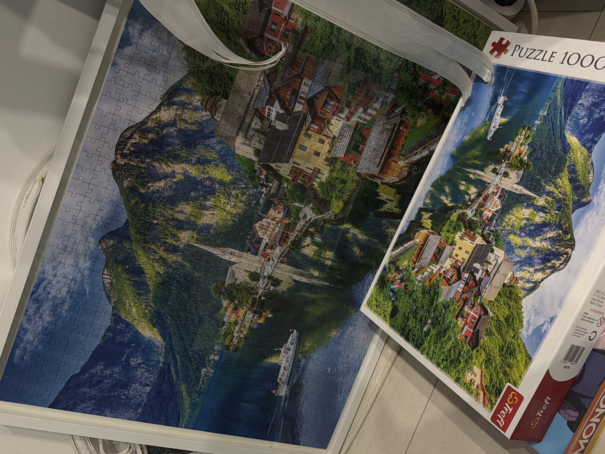 The image shows two puzzles featuring a scenic view of Hallstatt, Austria. The larger, framed puzzle, partially visible, displays the picturesque lakeside town with mountains in the background, a church steeple, and a boat on the water. The smaller puzzle, in its box, also depicts a similar scene of Hallstatt. The box has the brand "Trefl" and indicates "Puzzle 1000". The scene is indoors, with the puzzles placed on a light-colored wooden surface.