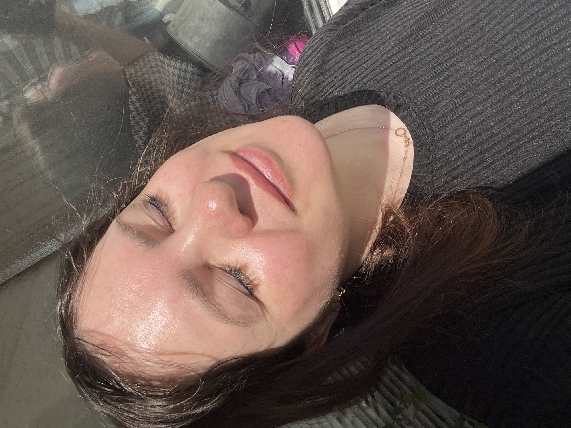 The image shows a close-up of a young woman with her eyes closed, basking in sunlight. Her face is tilted upwards, and her skin appears radiant, reflecting the natural light. She has long, dark brown hair that falls around her shoulders. She is wearing a black, ribbed shirt and a delicate gold necklace with a circular pendant adorned with small charms. The background is blurred, but suggests an outdoor setting, possibly near a window with a reflection of some objects and patterns. There is no indication of the location being in Noviy, Russia, or any other specific city.