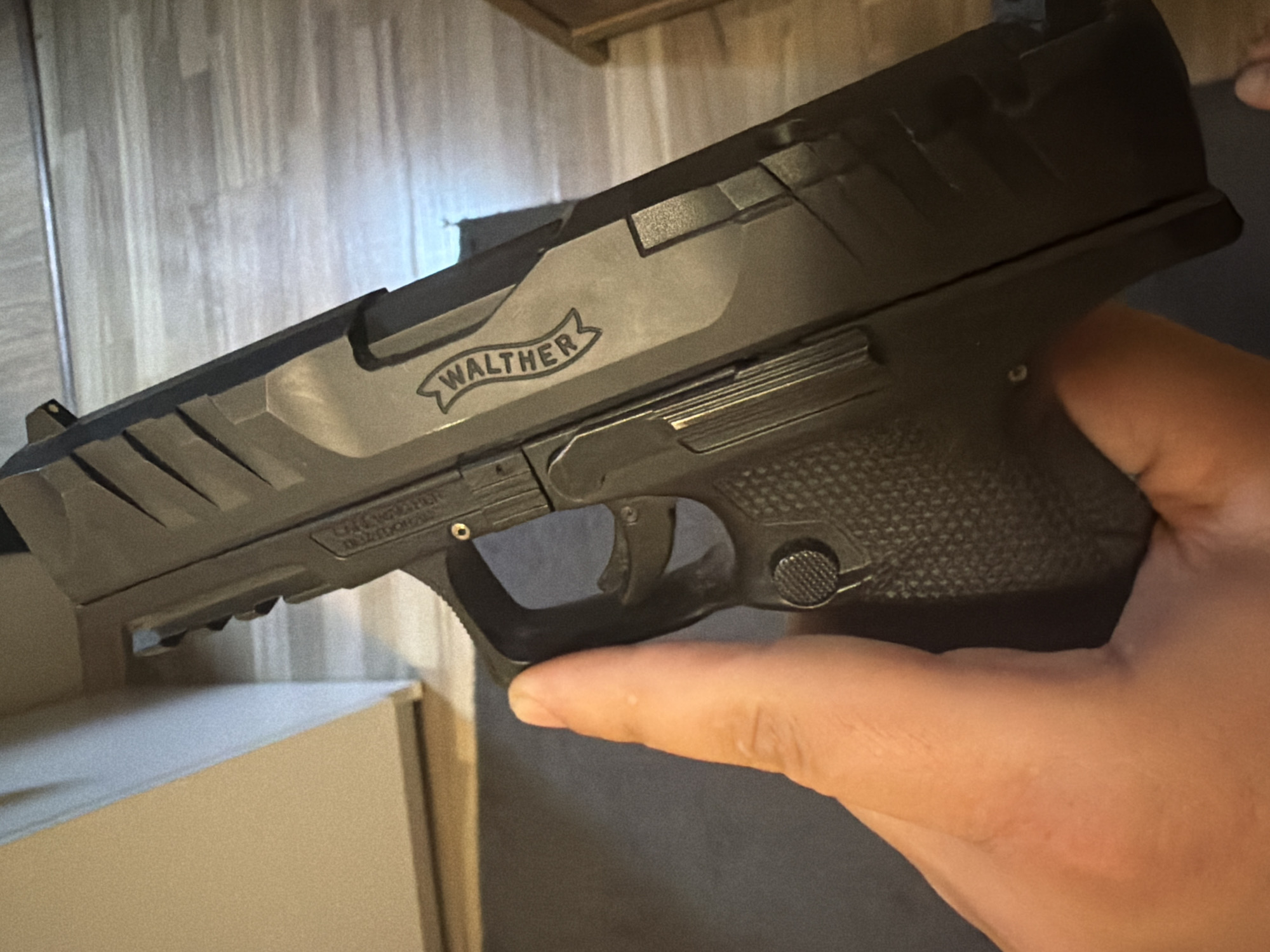 A close-up shot captures a person's hand holding a black semi-automatic pistol in an indoor setting. The pistol is angled, with its barrel pointing towards the upper left, while the right hand firmly grips the textured handle in the lower right of the frame.
The pistol features a dark, matte finish with distinctive aggressive serrations on the slide and a stippled or textured grip for enhanced handling. The brand name "WALTHER" is prominently engraved in a banner design on the left side of the slide. Below the Walther logo, smaller text is visible, partially reading "PDP COMPACT 9x19", indicating the specific model and caliber of the firearm. The user's trigger finger is safely positioned outside the trigger guard.
The background is softly blurred, revealing a light-colored wooden surface, possibly flooring or wall paneling, composed of vertical planks. A light-toned, rectangular object, resembling a box, is partially visible in the lower left corner of the background. The lighting is diffused, suggesting an indoor environment, with no discernible outdoor elements or specific time of day indicated. The image focuses primarily on the firearm and the hand holding it, without revealing further location-specific details.