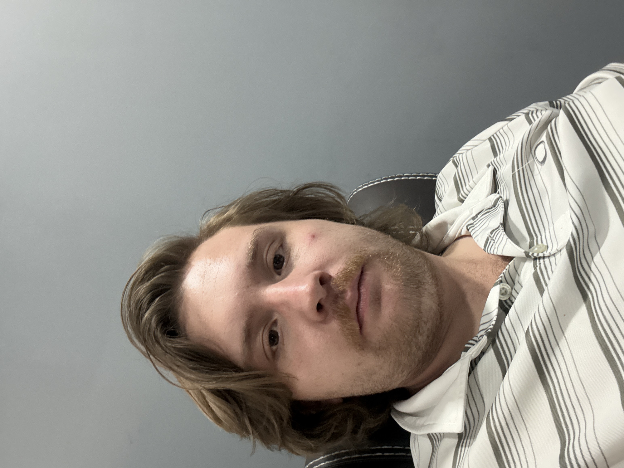 A close-up, slightly angled shot shows a fair-skinned man with light brown hair and a short, unkempt beard. He is wearing a light-colored striped shirt. The background is a solid gray wall. No specific details suggest a location in Cuautitlán Izcalli, Mexico.