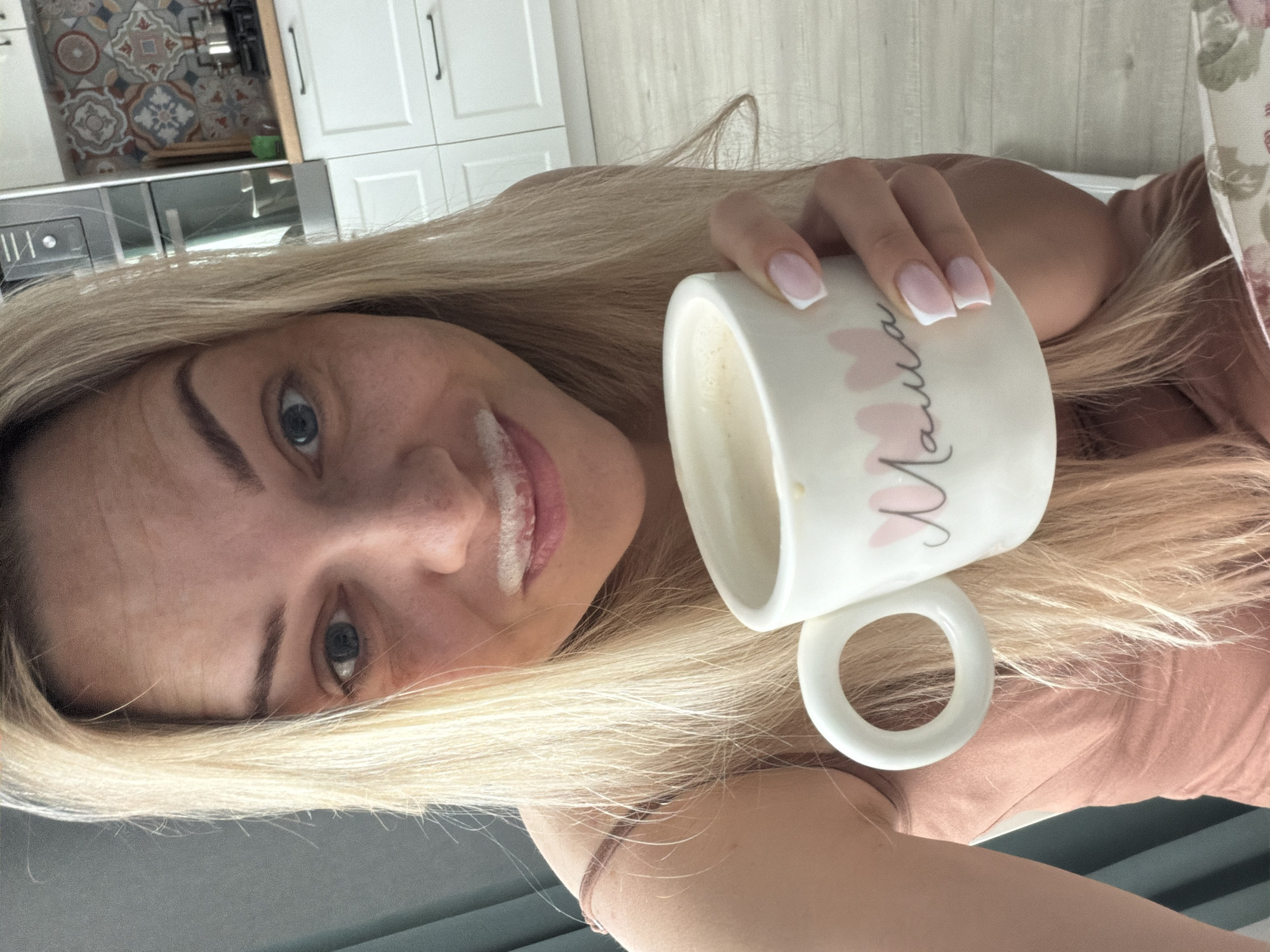A blonde-haired woman with fair skin holds a white mug with the word "Мама" (Mama) and pink heart images on it. There is a milk mustache on her upper lip. The mug contains a light-colored liquid, likely milk or a milk-based drink. The woman's nails are manicured with a French tip design. She is wearing a peach-colored top. The background shows a kitchen with white cabinets and decorative tile above a countertop. The image is rotated to the left.