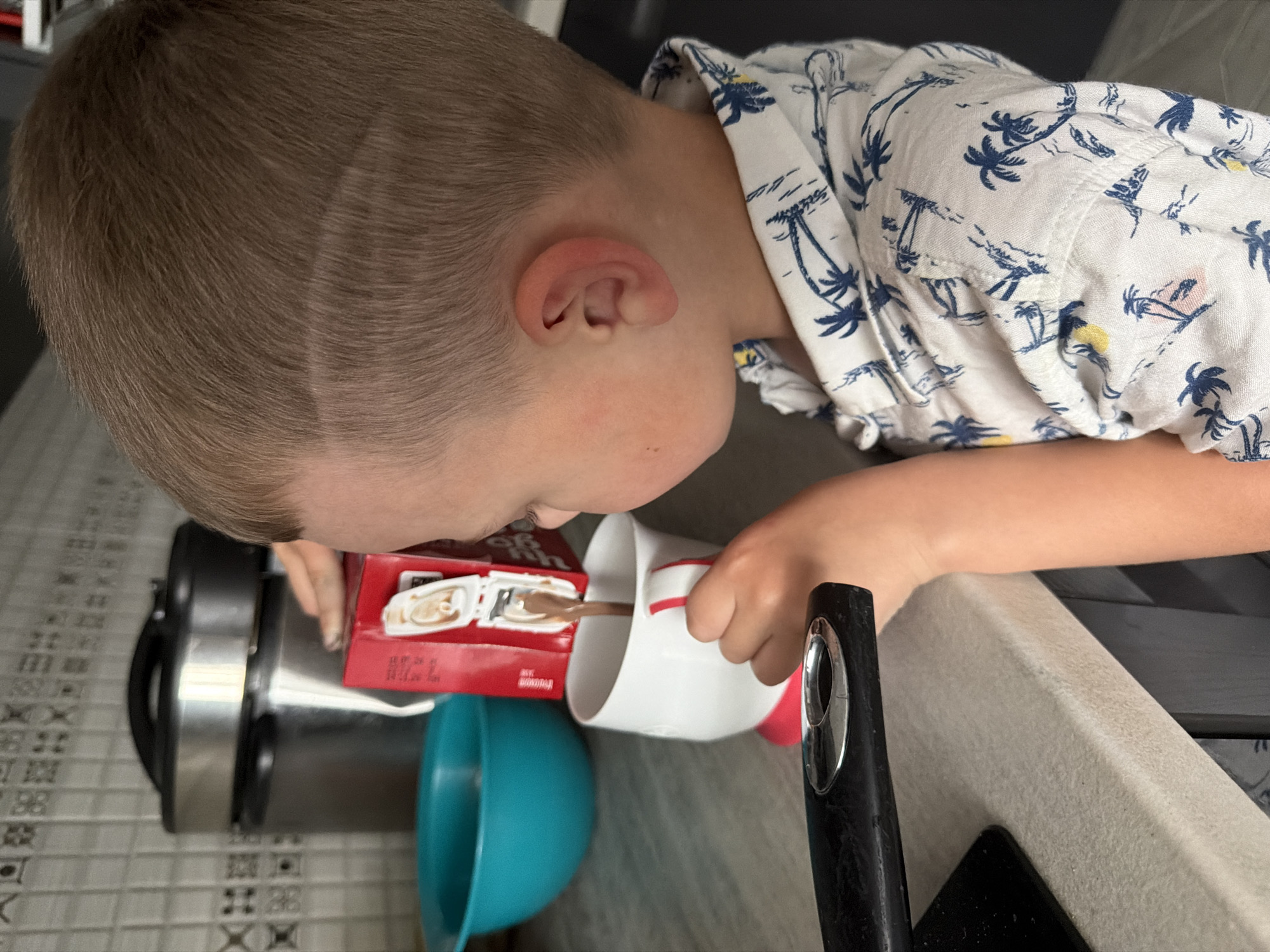The image shows a close-up, low-angle shot of a young child pouring a beverage from a small red box into a white mug. The child is wearing a white short-sleeved shirt with a blue palm tree print. Their hair is short and brown, with a subtle fade or design on the side. The child's left hand holds the red box, labeled with text that is partially visible, including "CHOCOLATE DRINK". Their right hand is holding the white mug, which has a red rim and appears to be mostly empty except for the liquid being poured. A spoon with a wooden handle is inside the mug, and the liquid is a light brown color.
In the background, a turquoise bowl is visible to the left of the mug. Behind the child and the mug, a dark, metallic object, possibly a kitchen appliance, can be seen. The setting appears to be a kitchen or dining area, with a countertop and tiled flooring visible. The overall lighting suggests it might be daytime. There are no specific cues about the weather. The image captures a moment of an everyday activity, with the child focused on pouring the drink.