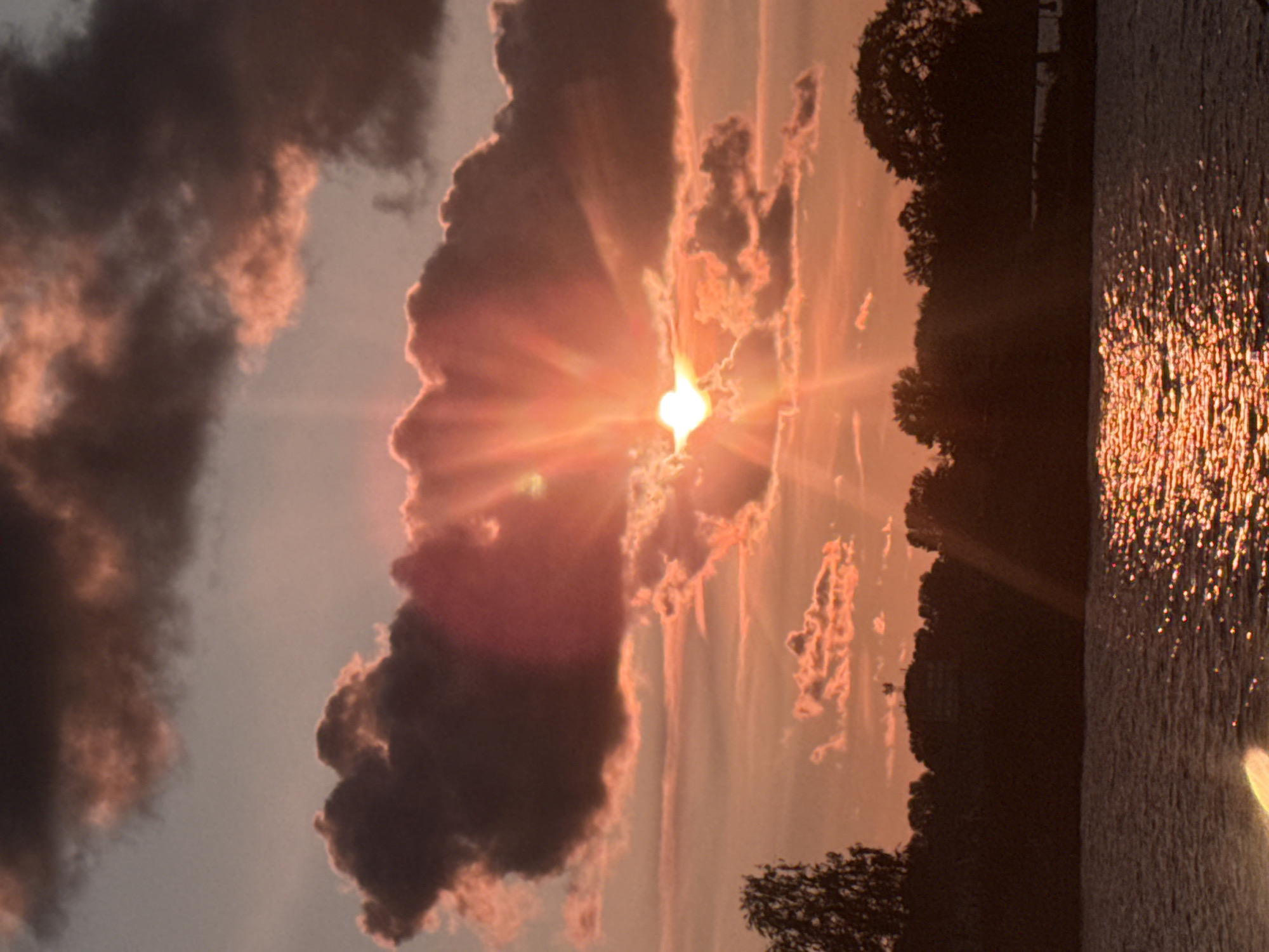 This is a photograph of a sunset over a body of water, likely a river or lake, in Riga, Latvia. The sun is partially obscured by clouds, casting a warm orange and pink glow across the sky. Rays of sunlight emanate from behind the sun, creating a dramatic effect. Silhouetted against the colorful sky is a treeline with a few buildings visible on a hill in the distance, one of which has an antenna or tower. The water in the foreground reflects the sunset, creating a shimmering effect. The overall atmosphere is tranquil and serene.