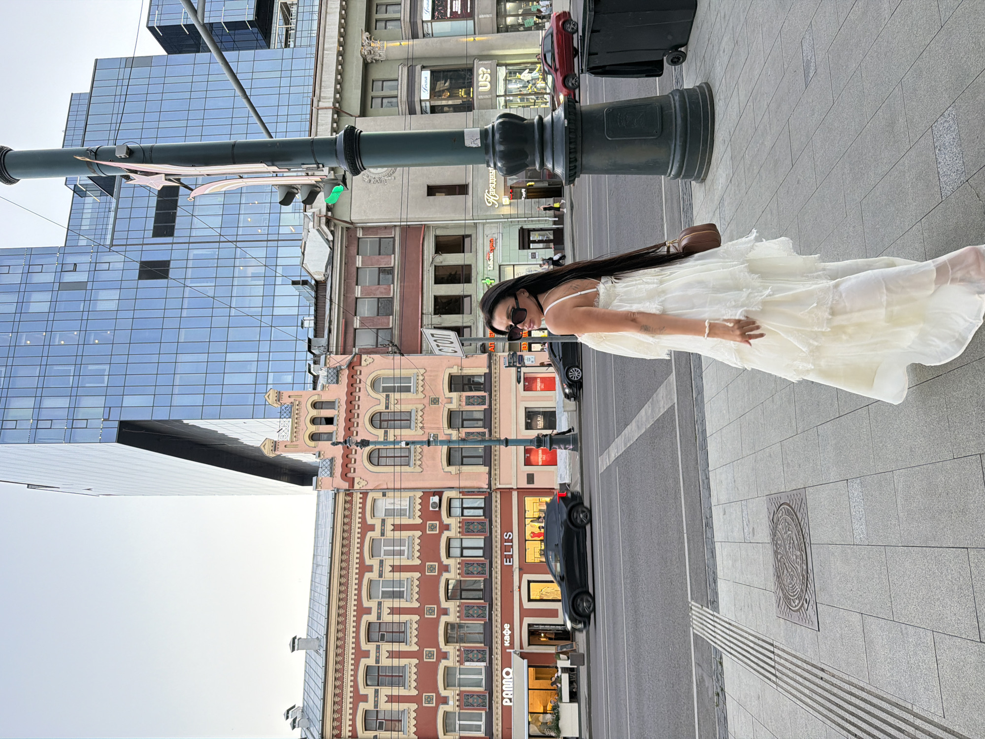 A woman with long dark hair and sunglasses is standing on a sidewalk in front of a building. She is wearing a white, ruffled, floor-length dress and carrying a brown purse. The scene is urban, with a modern glass building on the left and older, ornate buildings on the right. A street with cars and a traffic light is visible. The sky is overcast, suggesting it is daytime. There are signs on the buildings, including one that says "ELIS" and another that appears to be in Cyrillic. The overall mood is casual and relaxed.