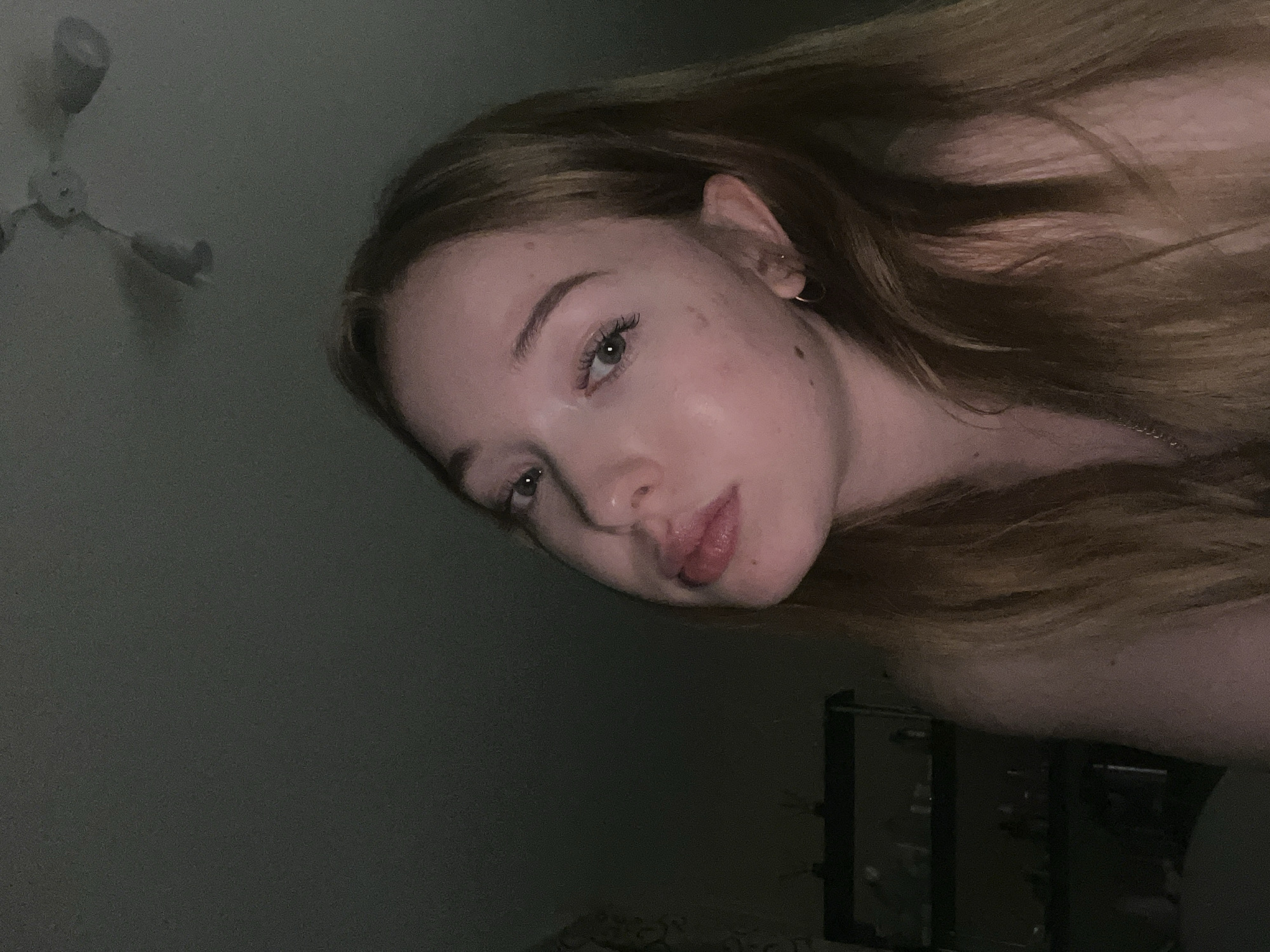 A dimly lit interior scene features a woman with long, light-brown hair. She is oriented at an angle to the camera. Her face is partially illuminated, revealing fair skin, light-colored eyes, and freckles. She wears a small earring in her left ear. The background is dark, with a ceiling fan visible in the upper left corner, and a dark object in the lower right that could be a piece of furniture. The location in the city of Chaikovsky, Russia is not discernible from the image.