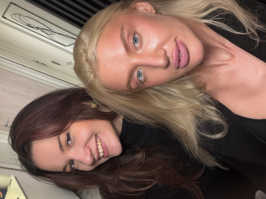 Two women pose closely for a selfie. The woman on top has blonde hair, fair skin, and light blue eyes. Her lips are a light pink color. The woman below her has shoulder-length brown hair with red highlights. She has brown eyes and is smiling, showing her teeth. Both women are wearing black tops. The background includes a white door and what appears to be a black slatted ceiling.