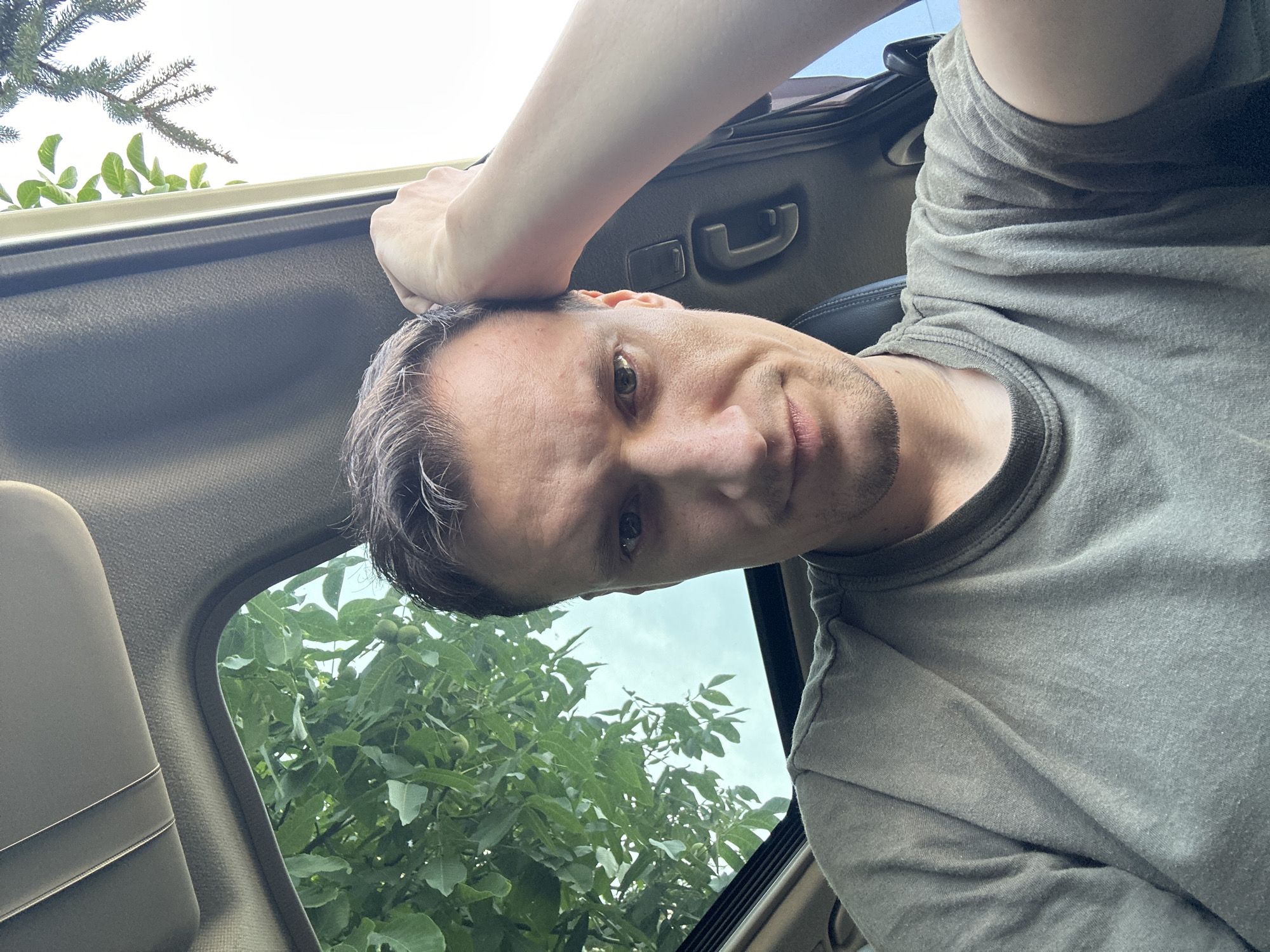 A man with short brown hair and stubble on his chin rests his arm on his head as he looks directly at the camera. He is wearing a plain olive-green t-shirt. The man is inside a vehicle, with the sunroof open, revealing green leaves and branches from a tree above. The overall lighting suggests it is daytime. The image was taken in Shatava, Ukraine.