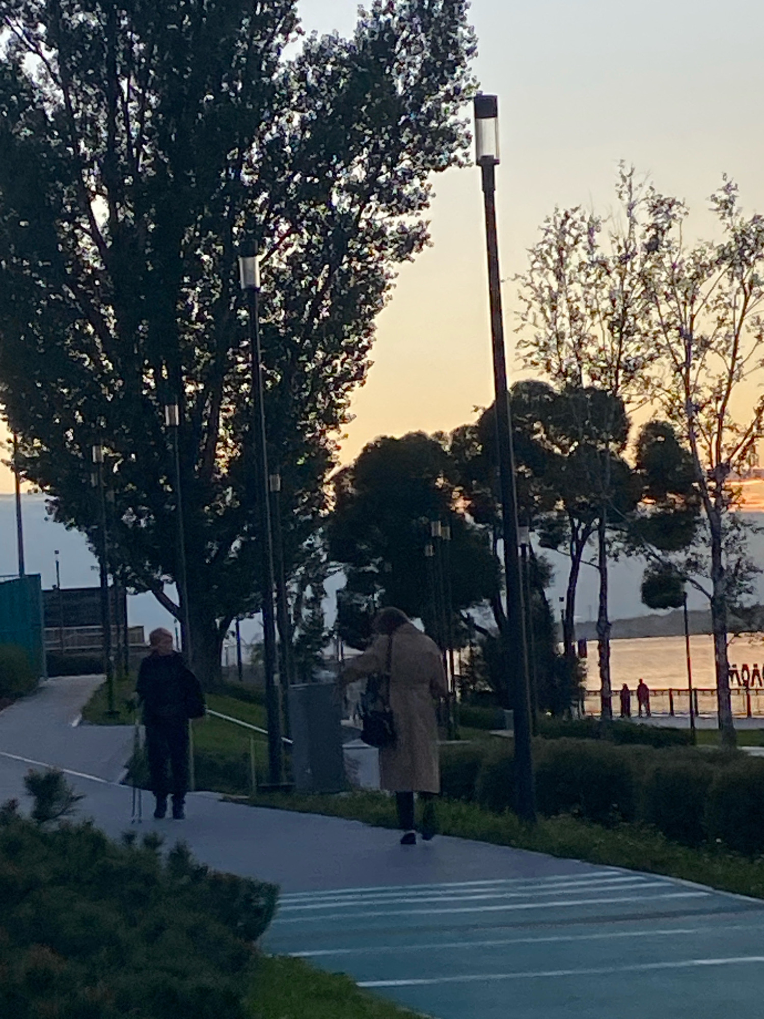 The image depicts an outdoor scene at dusk, likely in Perm, Russia, given the presence of typical urban park infrastructure. In the foreground, a paved pathway with distinct white markings curves to the right. To the left, dense green foliage borders the path. Further back, two individuals are walking on the path. The person on the left, dressed in dark clothing, appears to be using a cane. The person on the right, wearing a light-colored trench coat and dark pants, carries a bag over their shoulder.
Behind the path, several tall trees with dark foliage are visible, interspersed with streetlights. In the middle ground, a large trash receptacle is positioned near the pathway. Beyond the trees and pathway, a body of water, possibly the Kama River, is visible, reflecting the warm hues of the setting sun. Silhouetted figures can be seen along the waterfront railing in the distance. The overall ambiance is serene, suggesting the end of a day in an urban park setting.
