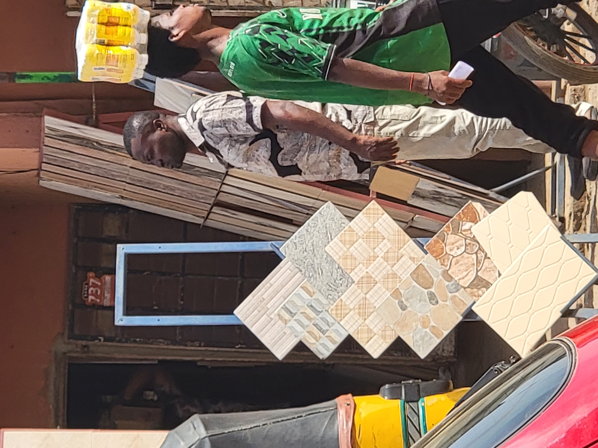 The image shows two men standing near a pile of floor tiles. One man is wearing a green shirt and black pants and is holding a small white object. The other man is wearing a patterned shirt and light-colored pants. The tiles are displayed in front of a building. A red sign with the number 737 is affixed to the building's brickwork. A motorcycle and a red vehicle are also visible in the foreground. Based on the user's prompt, the location is in the city of Maiburiji, Nigeria.