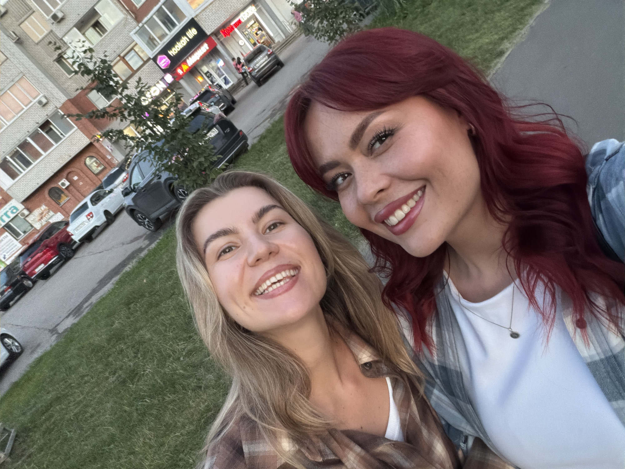Two young women are captured in a selfie on a street in Tolyatti, Russia. The woman on the left has blonde hair and is wearing a plaid shirt over a white top. The woman on the right has red hair and is wearing a white top with a plaid shirt. They are both smiling at the camera. In the background, there is a street with parked cars, including a white sedan, a red sedan, and a dark SUV. There is also a multi-story building with shops on the ground floor. The building is brick and has several windows. The shops have signs, one of which reads "hookah life". The overall environment appears to be an urban setting.