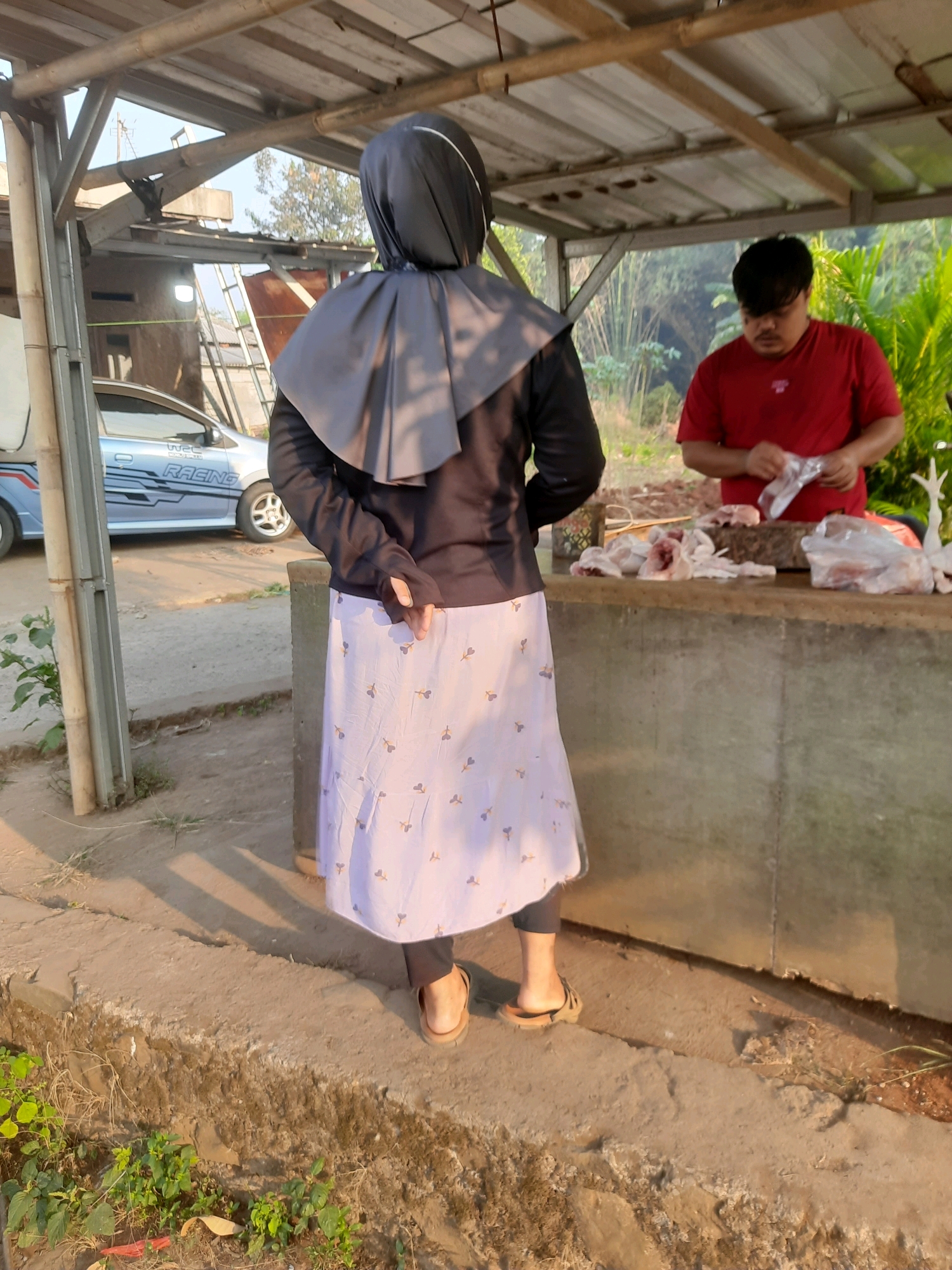 In Gandoang, Indonesia, a person wearing a black hijab and a light-colored skirt with a floral pattern stands facing away from the camera under a simple wooden structure. To the right, a man in a red t-shirt is seen from behind, actively handling plastic-wrapped meat at a concrete counter. The environment appears to be an outdoor market or food stall, with a light blue car parked in the background. The scene suggests a transaction or preparation of food in a semi-rural setting.