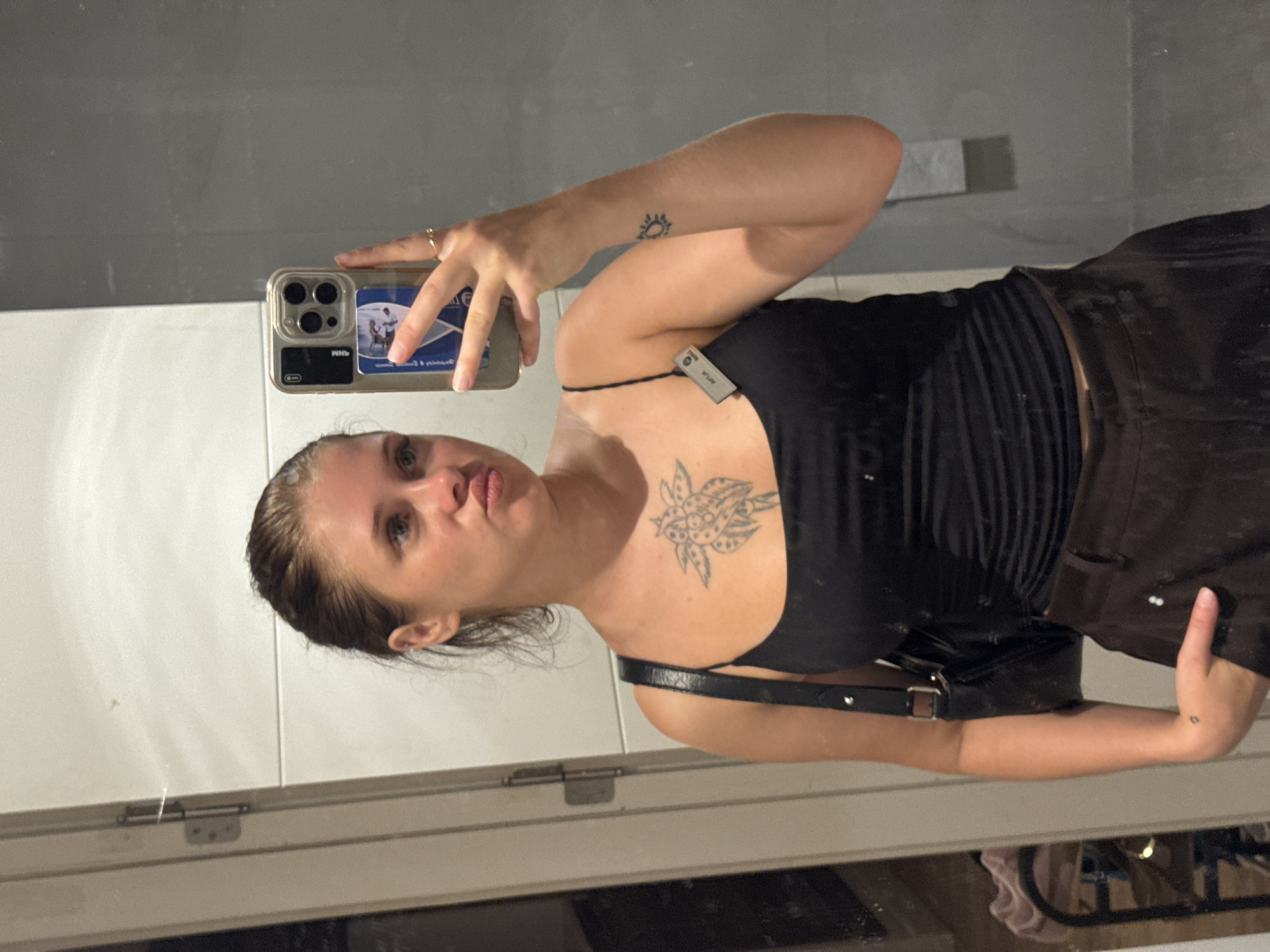 A young woman is reflected in a mirror, holding a smartphone up to take a selfie. She has her hair pulled back in a bun and is wearing a black tank top and brown pants. A black purse is slung over her shoulder. She has tattoos on her chest and forearm. The background appears to be a bathroom, with tiled walls and a door. There is no indication of the city of Aksu, Turkey in the image.