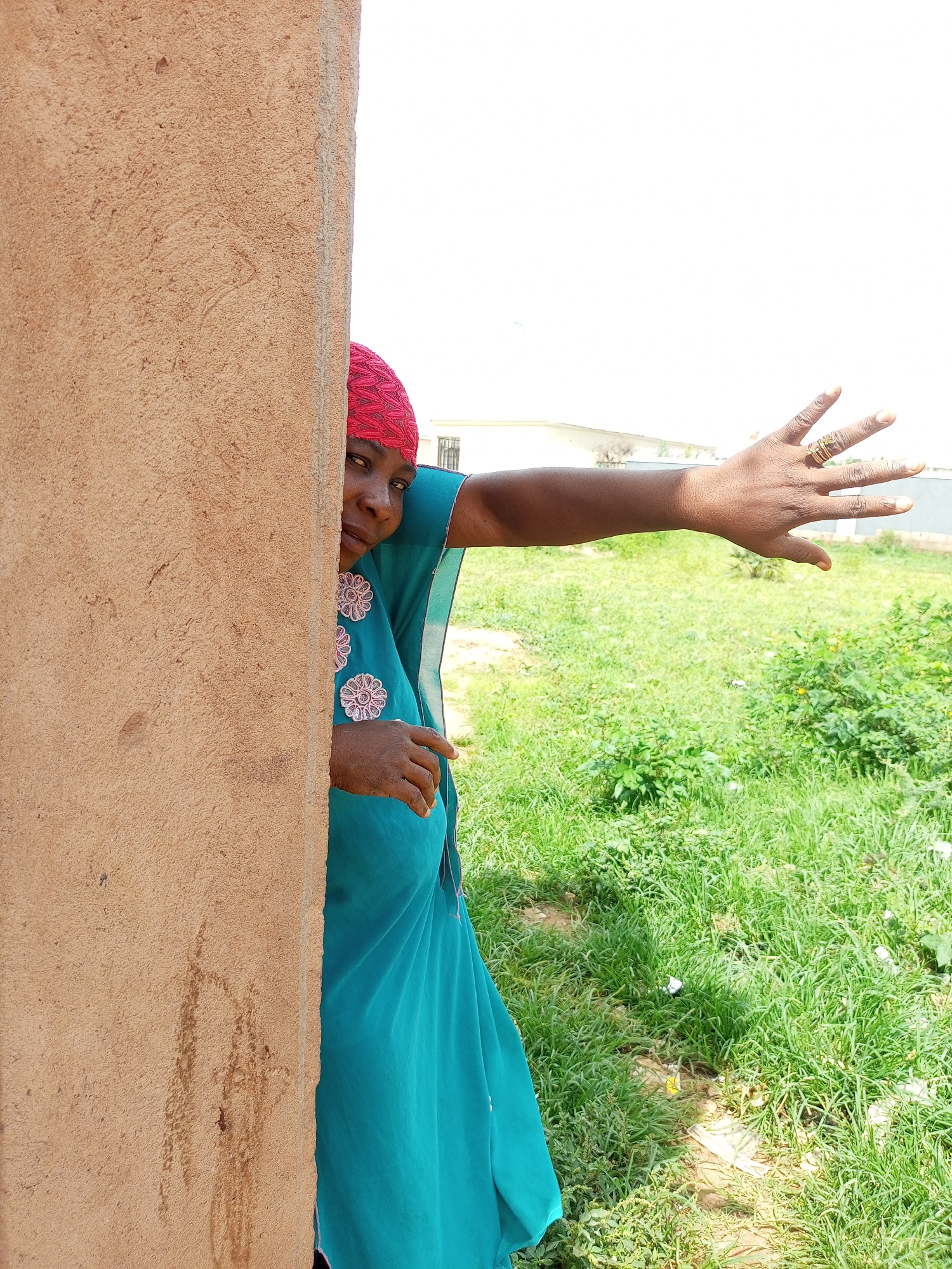 In the city of Tempure, Nigeria, a woman with a red headscarf and a teal dress with embroidered flowers peeks from behind a textured tan wall. She extends her right arm, palm open, towards the camera. Her hand displays multiple rings on two fingers. The background is a bright, overcast sky and a field of green grass dotted with bushes and some scattered white debris. Some buildings are visible in the distance.