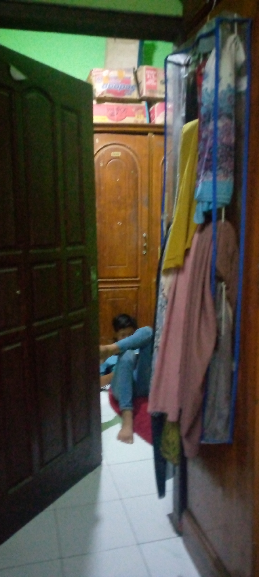 In Sumberwono, Indonesia, a person in blue jeans and a light blue shirt sits on a red mat in a room. To the left, a dark brown wooden door stands ajar. Further into the room, a tall wooden door with an arched top is visible. To the right of the wooden door, a blue garment rack holds several pieces of clothing, including a yellow garment, a pink garment, and a blue and white patterned garment. In the background, several cardboard boxes are stacked on a shelf. The floor is tiled with a light-colored tile.