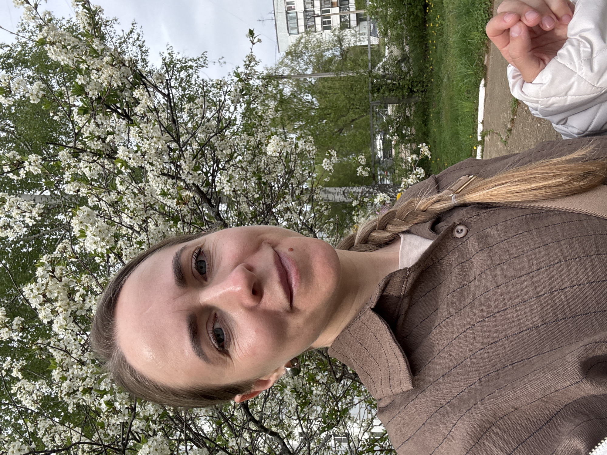 A woman with light skin and blonde hair braided and secured with a barrette is the primary subject of the photograph. She is wearing a brown, striped linen shirt and faces the camera with a slight smile. Her gaze is direct.
The background features a flowering tree with white blossoms, indicating springtime. Behind the tree, a blurred urban landscape includes a multi-story building and green foliage. A white jacket is seen at the right edge of the frame.
The setting is outdoors in Chaikovsky, Russia. The photo appears to be a self-portrait or taken at close range by another individual.
