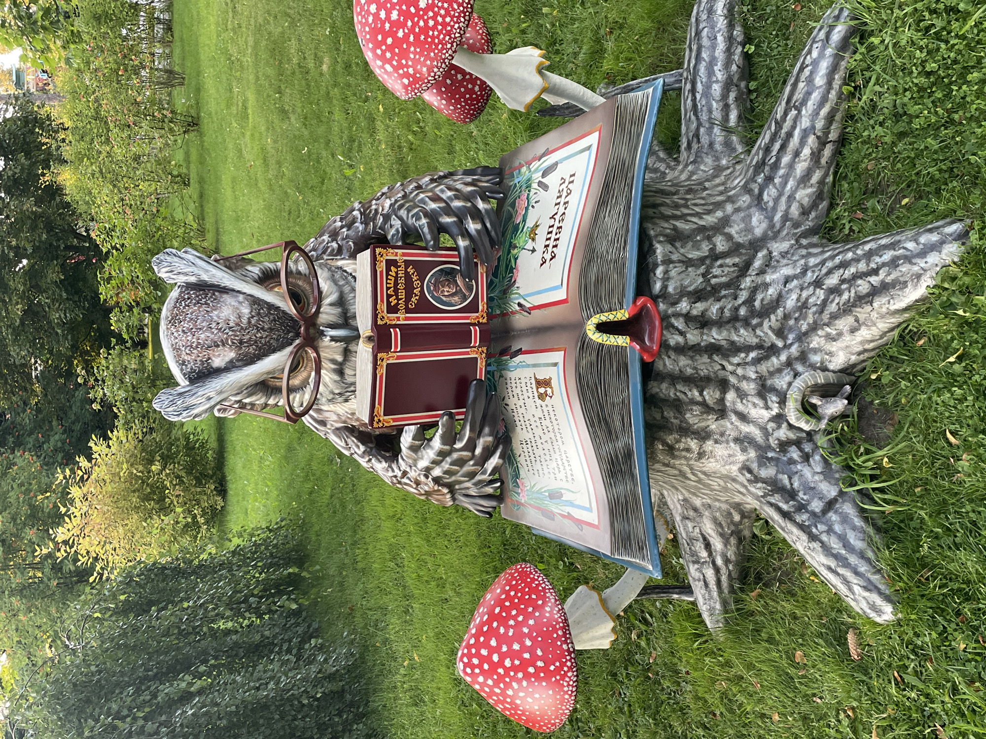 A whimsical owl sculpture wearing glasses sits on a tree stump in a park, reading an open book. The owl, made of sculpted material, is detailed with feathers and large eyes. It holds a smaller closed book in one wing. The open book displays illustrated pages with Russian text. Two large, red and white spotted mushrooms, resembling Amanita muscaria, flank the tree stump. The sculpture is situated on a grassy lawn, surrounded by trees and bushes. The background suggests a well-maintained park or garden. This scene is located in Saint Petersburg, Russia, specifically in the Pavlovsk Park.