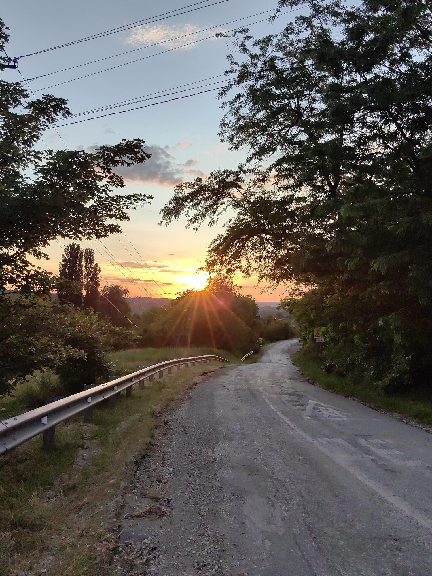 A dirt road curves uphill, bordered by a metal guardrail on the left and dense foliage on the right. The sky is a pale blue with wisps of clouds catching the warm glow of a setting sun. Sunbeams dramatically fan out from behind a cluster of trees on the right, illuminating the road and surrounding landscape with an orange hue. Power lines are visible stretching across the sky, strung between utility poles on the left. The road appears worn and uneven, with patches of gravel and some visible potholes. The overall scene suggests a rural or semi-rural setting in the Ароматновское сельское поселение, Ukraine, during the golden hour.