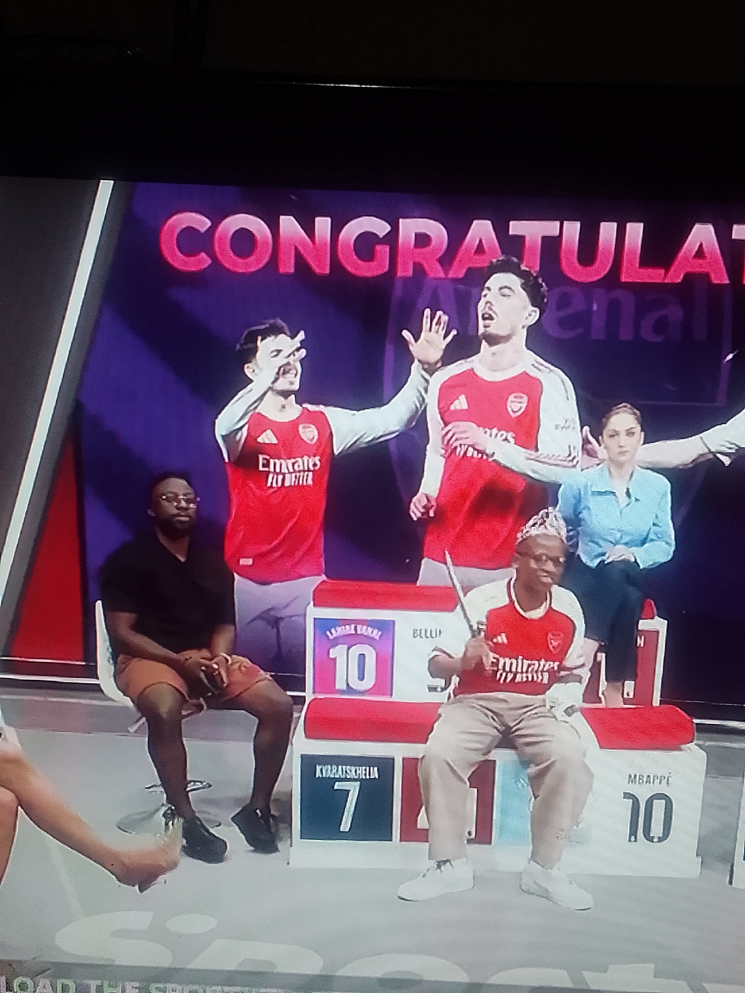Here is a description of the image:
The image shows a scene from a television show set. In the background, a large screen displays the word "CONGRATULATIONS Arsenal". Two images of soccer players wearing Arsenal jerseys are visible on the screen.
In the foreground, there are four people. On the left, a man with dark skin, wearing a black shirt and brown shorts, sits in a chair. To his right, a man with dark skin, also wearing an Arsenal jersey, sits on a podium holding a small sword. The podium has numbers and names written on it, including "10 LAHIRE VANAL", "BELLUP", "7 KVARATSKHELIA", and "10 MBAPPE". A woman wearing a light blue shirt sits behind the podium. Another woman is visible in the lower left.