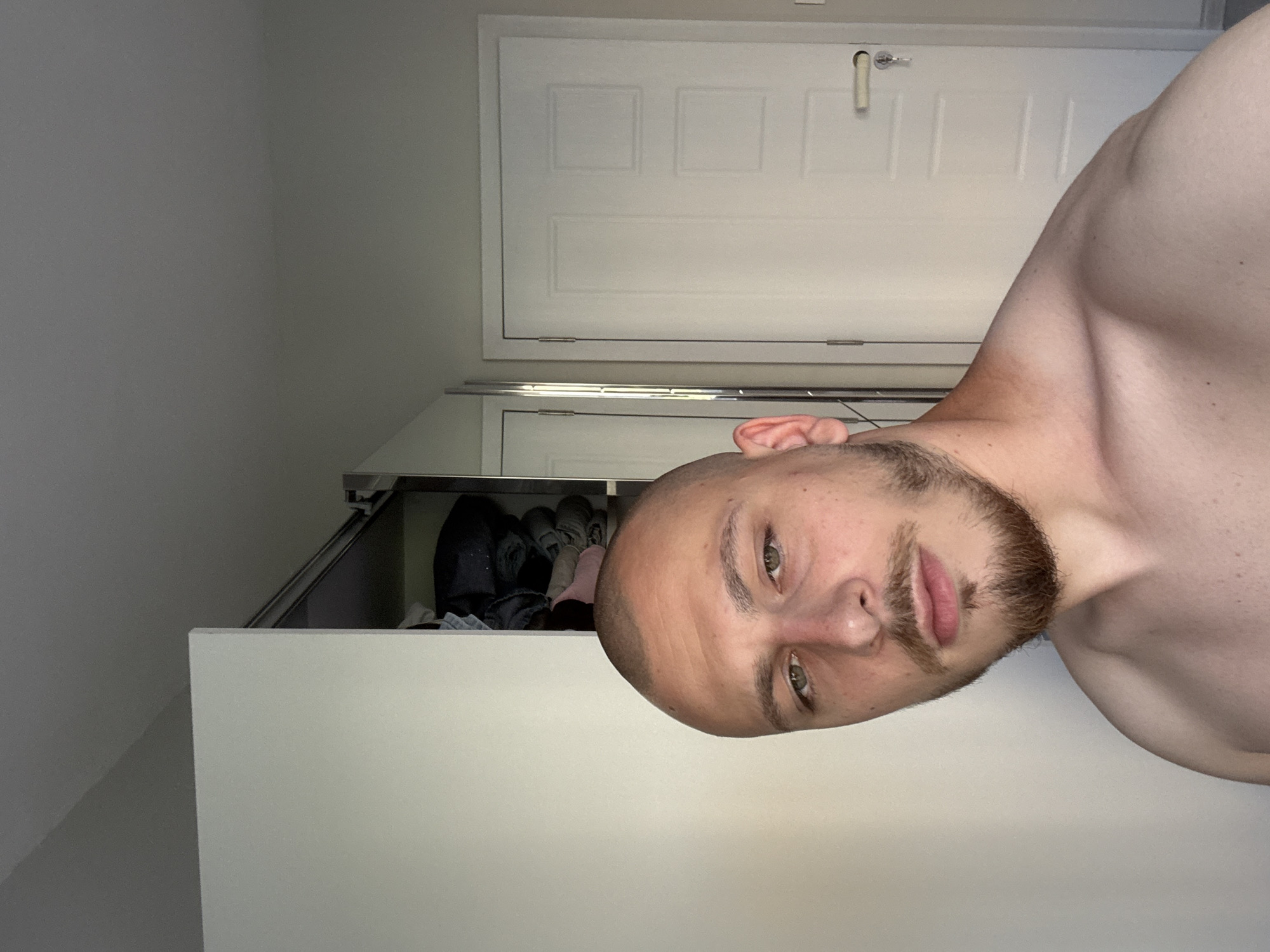 A young adult male, appearing in his twenties, with a shaved head and a short, neatly trimmed brown beard and mustache, is depicted shirtless, facing the camera directly with a neutral expression. His bare shoulders and upper chest are visible. The setting is an indoor residential space in Stara Zagora, Bulgaria. In the background to the left, a white wardrobe door is partially open, revealing stacked clothing items inside. To the right, a white interior door featuring three rectangular panels, a silver handle, and an inserted key is visible. The room has light-colored walls and appears evenly lit.