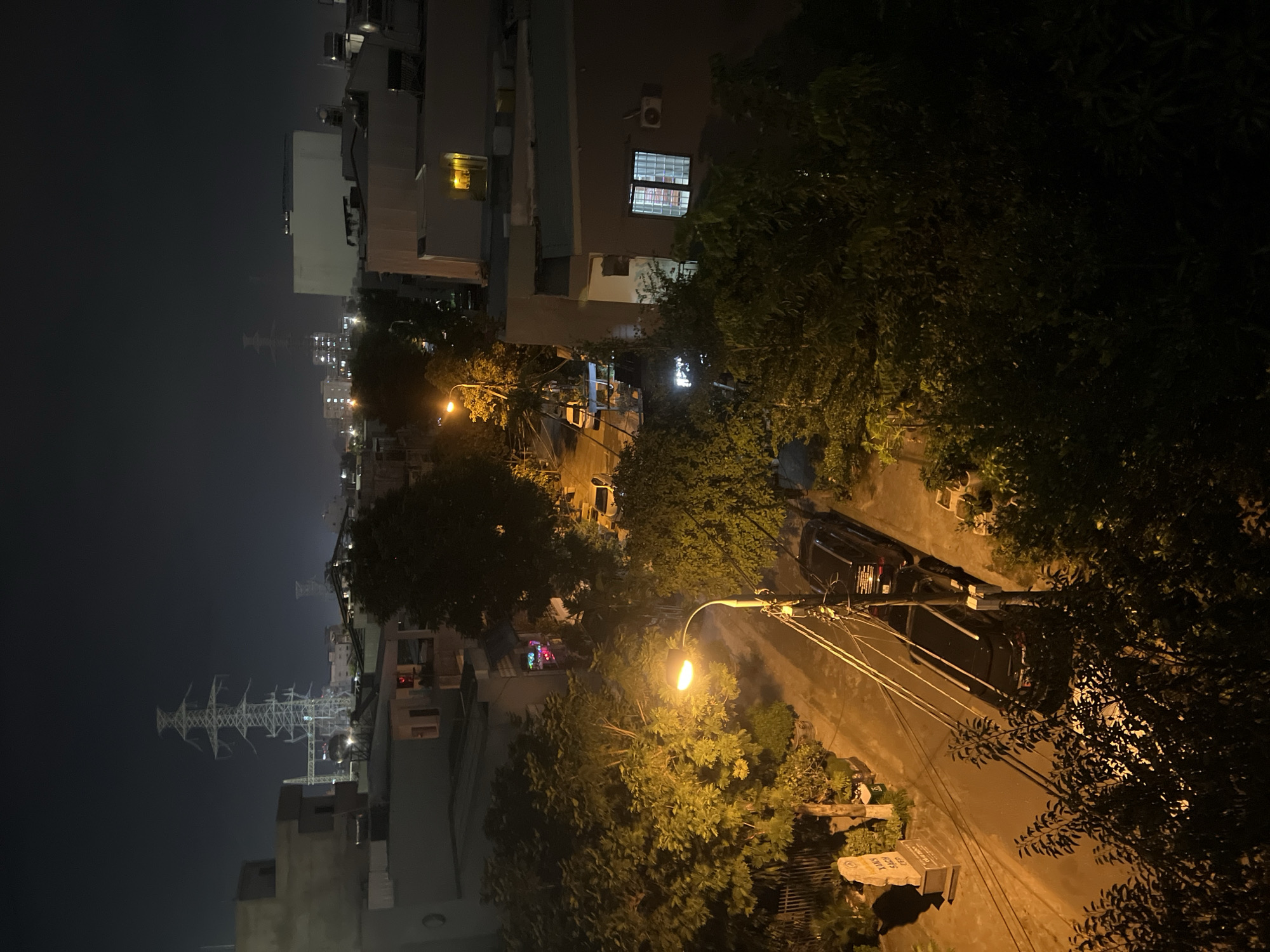 This image captures a nighttime view of a street in the city of Da Nang, Vietnam, likely a residential area. The foreground is dominated by dark, leafy trees, with some branches illuminated by the warm glow of a streetlamp. A black SUV is parked on the right side of the asphalt road, partially obscured by foliage. Further down the street, another black vehicle, possibly also an SUV, is visible. A streetlamp with an orange light illuminates a section of the road and sidewalk on the left. Beside the sidewalk, a stone monument or marker stands with text inscribed on it, casting a shadow. Behind the trees and street, a multi-story building with several windows is present, some lit from within. In the background, the cityscape unfolds with more buildings of varying heights and architectural styles. Prominent in the distance are towering electricity pylons, a common sight in urban and semi-urban areas. The sky is dark, indicating it is nighttime, with a slight haze or light pollution contributing to the overall ambiance. There are no people visible in the frame. The scene is quiet and appears to be a typical residential street at night, with the electric infrastructure of the city serving as a backdrop.