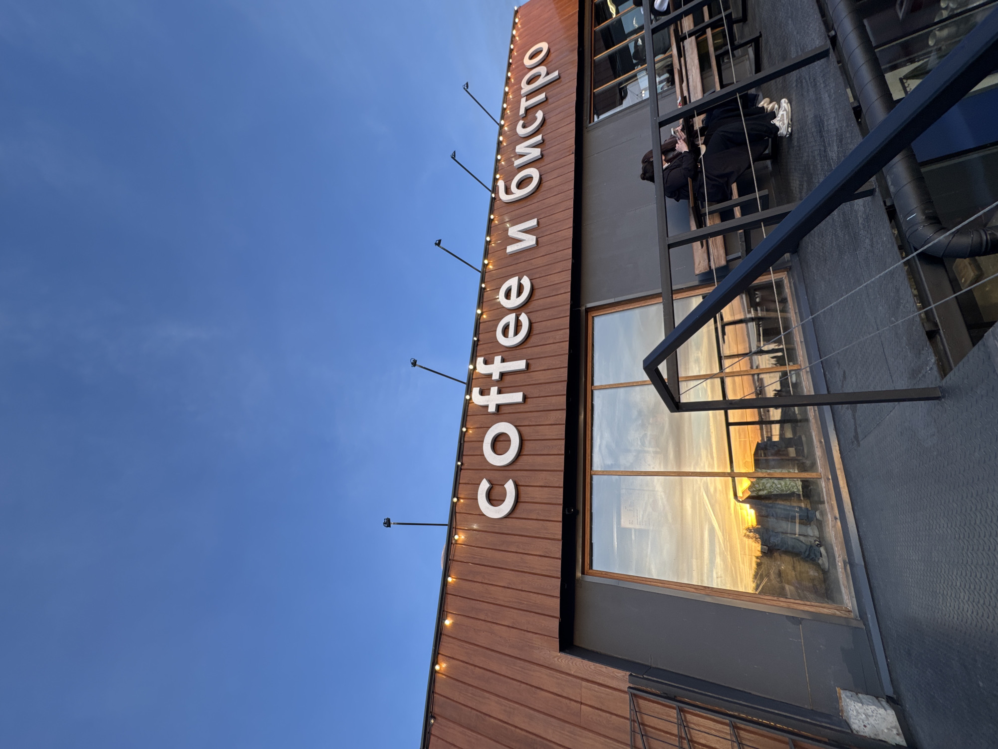 Here is a description of the image:
The image depicts the exterior of a building with a sign in Cyrillic and English that reads "Coffee и Бистро" ("Coffee and Bistro"). The building has a wooden facade with vertical paneling on the left side of the sign. String lights are along the edge of the wooden paneling. The building appears to have multiple levels with outdoor seating, as visible on a balcony with metal railings. Two individuals are visible on the balcony, seated on a bench. Their clothing is dark. Large windows reflect the scene of a sunset. The sky is clear and blue. The photograph is taken from a low angle, emphasizing the height of the building and the sky.