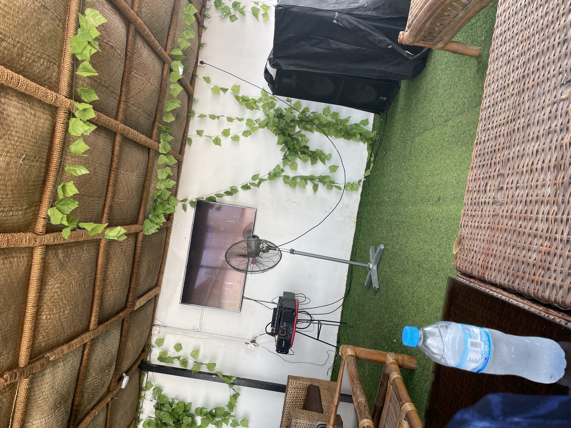 The image depicts an outdoor seating area with a thatched roof and artificial turf flooring, adorned with artificial ivy vines. In the background, a wall features a large flat-screen television mounted above a portable audio amplifier and speaker system. A standing fan is positioned to the right of the equipment. Further to the right, a large black speaker cabinet is covered with a protective cloth. In the foreground, a bottle of water rests on a wicker table, with glimpses of wicker seating. The scene appears to be a recreational or entertainment space, possibly a bar or lounge, located in Jauro Yinu, Nigeria. There are no people visible in the image.