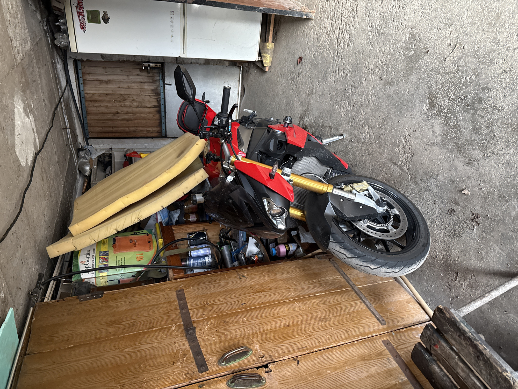 The image depicts a cluttered storage area in what appears to be a garage or shed. A red and black BMW motorcycle is prominently positioned, partially obscured by various items. In the background, there is a white appliance, potentially a refrigerator, stacked above a wooden door or hatch. A wooden chest or storage unit is in the foreground, covered with tools, cans, and other miscellaneous objects. Additionally, some pieces of yellow foam are also on top of the unit. The lighting is somewhat dim, and the overall impression is that of a cramped and disorganized space. The location is stated to be in Buhovo, Bulgaria.
