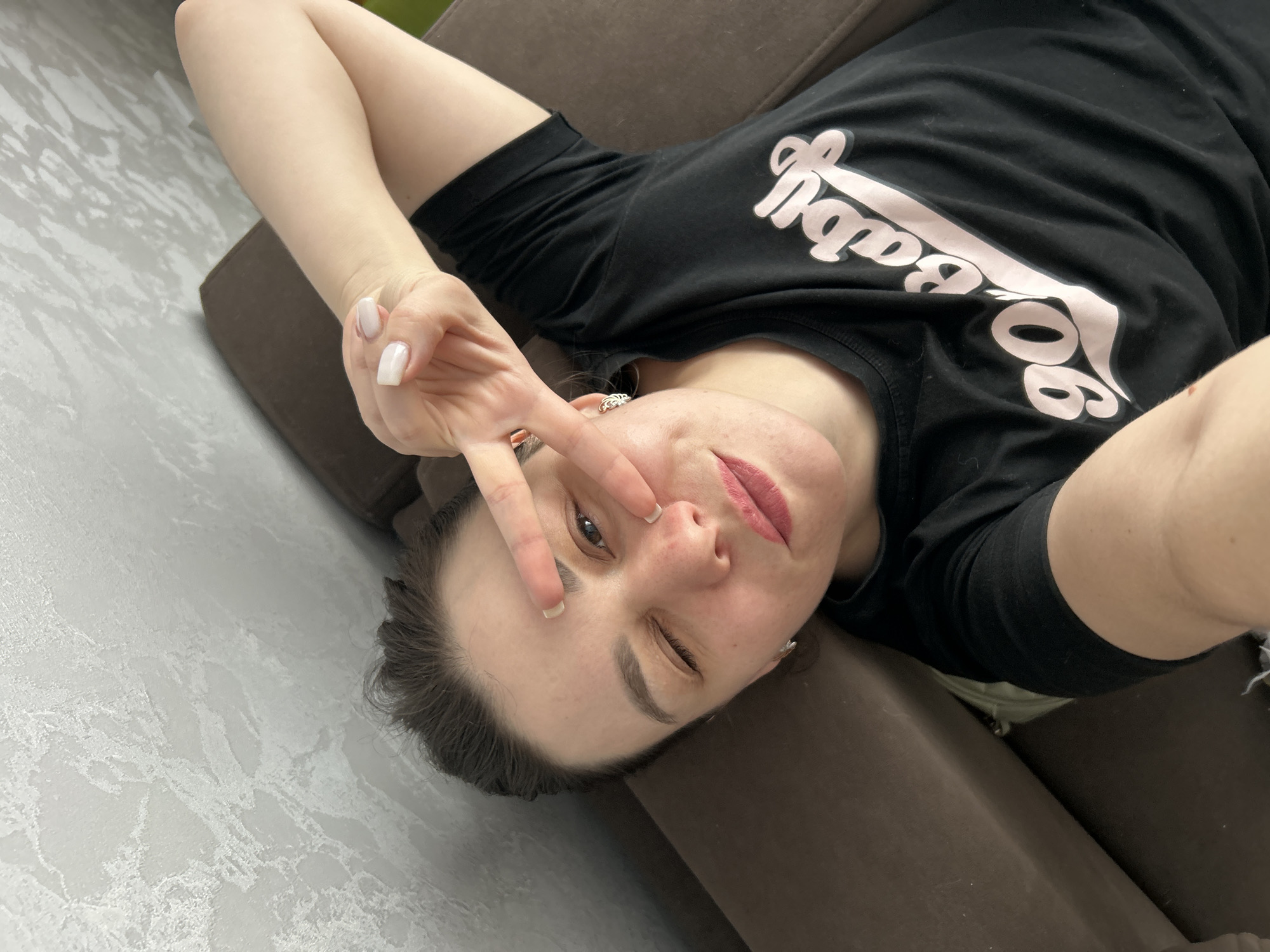 A woman is lying on a brown upholstered sofa in Chaikovsky, Russia. She is wearing a black t-shirt with pink lettering. She is winking with her right eye and holding up a peace sign with her left hand. Her nails are painted white. The background consists of a textured gray and white wall.