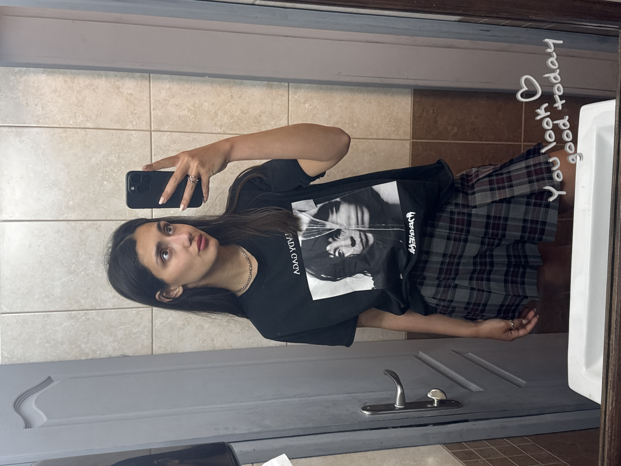 A young woman stands in a bathroom, reflected in a mirror. She is holding a phone in her right hand, presumably taking the picture. She is wearing a black t-shirt with a black and white print and a plaid skirt. The bathroom features light-colored tiled walls, a grey door with a silver handle, and a white sink. On the upper right side of the mirror, someone has written "You look good today" with a heart. The location is likely a residential or commercial bathroom, but the specific city of Bar, Montenegro, is not discernible from the image.