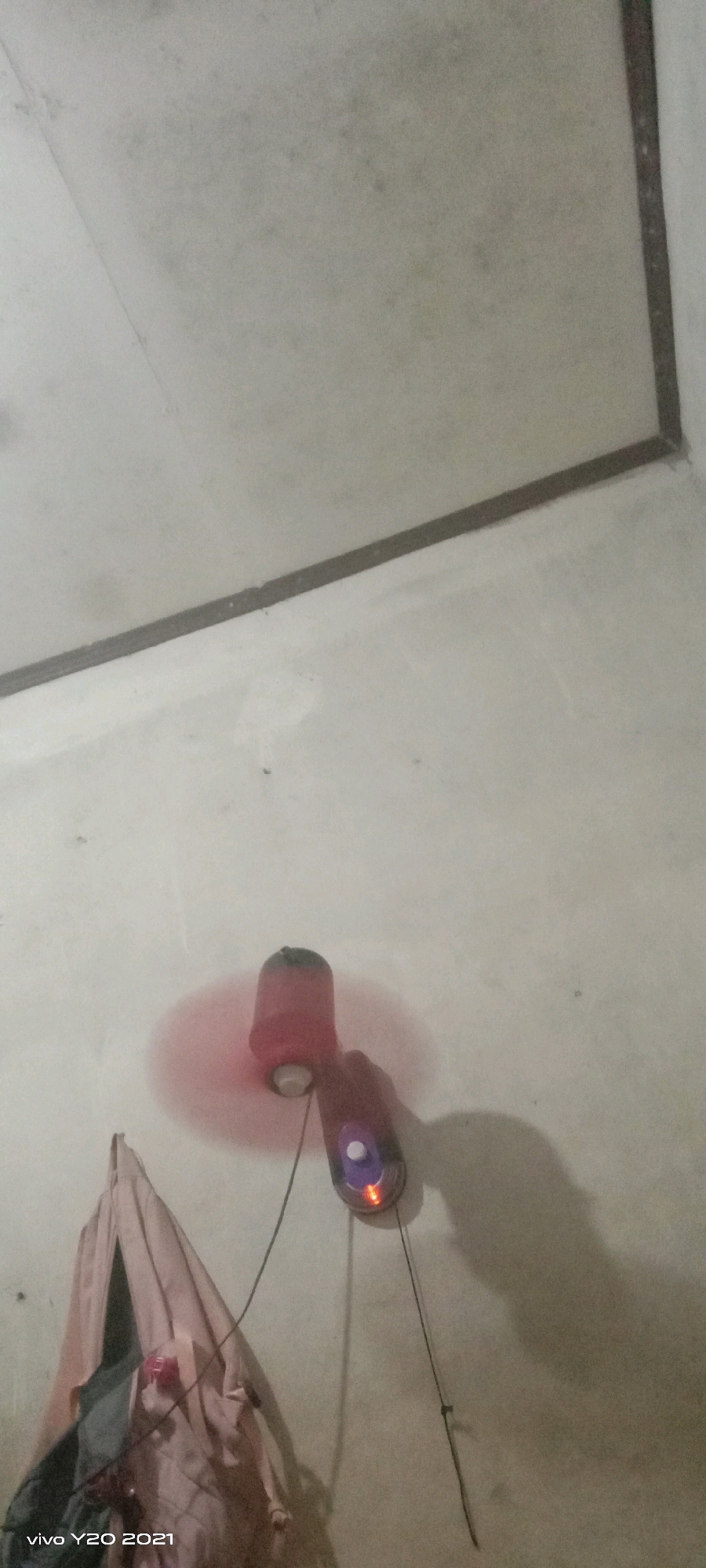 In an indoor scene, a small red fan with a dark top is mounted on a white wall. The fan is powered on, as indicated by the slight blur of its blades and the red glow around it. To the right of the fan, a black cord hangs down, attached to a dark object with a red light and a white button. A folded piece of light-colored fabric, possibly clothing or a cloth, hangs on the wall to the left of the fan. The wall appears somewhat textured and has a few small dark specks. The ceiling above is off-white and has a dark border along the top and right edges, suggesting a corner. A watermark in the lower-left corner reads "vivo Y20 2021".