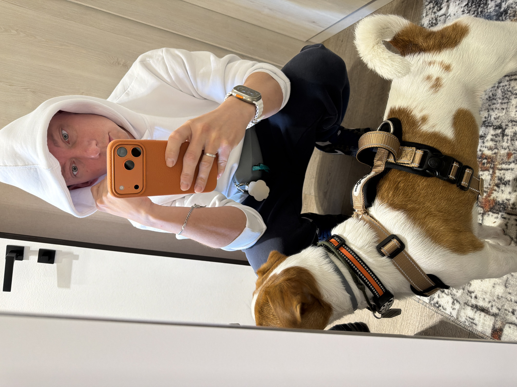 A person wearing a white hoodie and dark pants is taking a mirror selfie in a room with light-colored wooden walls and a patterned rug. The person is squatting, holding an orange smartphone to take the picture. A Jack Russell Terrier wearing a harness is standing in front of the person, looking towards the phone. The room appears to be an indoor space, possibly a dressing room or hallway, with a white door and black door handles visible on the left. The image does not provide enough information to determine the specific location within Moscow, Russia.