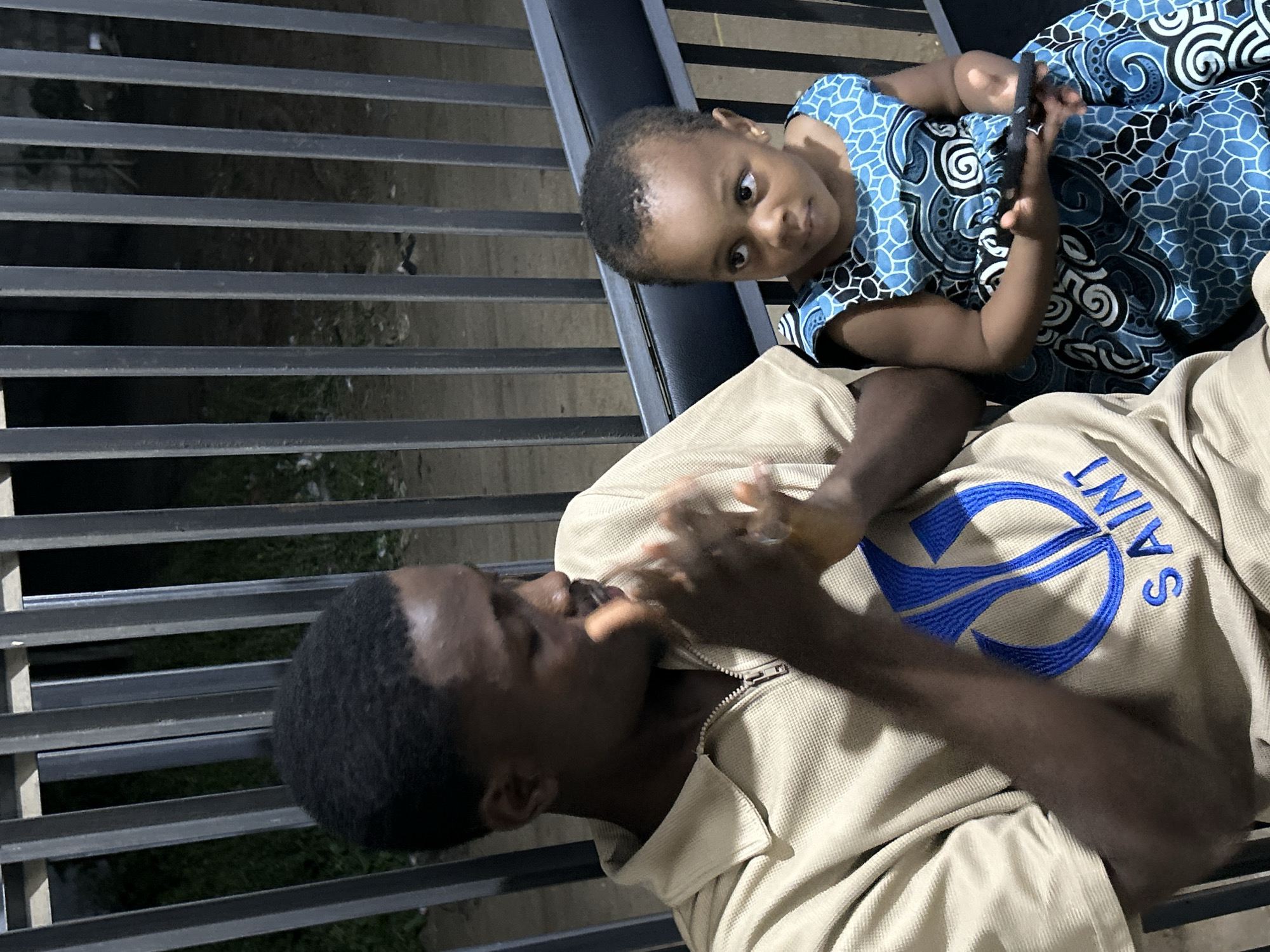 A man and a baby are together on what appears to be a bench or seating area. The man is African, dark-skinned, and wearing a tan jacket or sweatshirt with the word "SAINT" and an emblem on it in blue. He is leaning slightly back and appears to be putting something in his mouth. The baby, also dark-skinned, is wearing a blue and white patterned dress and is looking upwards, holding a dark-colored smartphone or similar device. They are positioned against a backdrop of metal bars, likely part of the seating area's design, through which greenery and possibly a sidewalk are visible. The location is inferred to be Ihiagwa, Nigeria, based on the prompt.