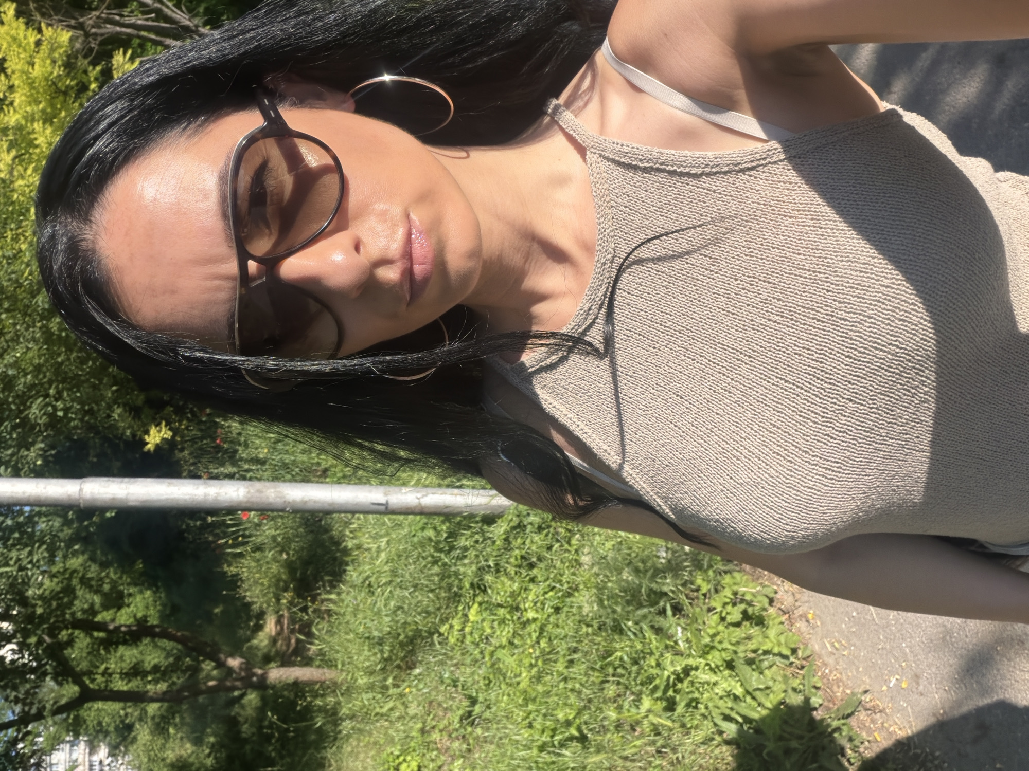 In an outdoor urban setting in Pleven, Bulgaria, a woman with long black hair and sunglasses is captured in a selfie. She is wearing a beige knitted tank top and large hoop earrings. The background features a paved path, abundant green foliage with some trees and red wildflowers, and a light-colored pole, likely a lamppost. The lighting suggests it is daytime, with shadows cast on the path.