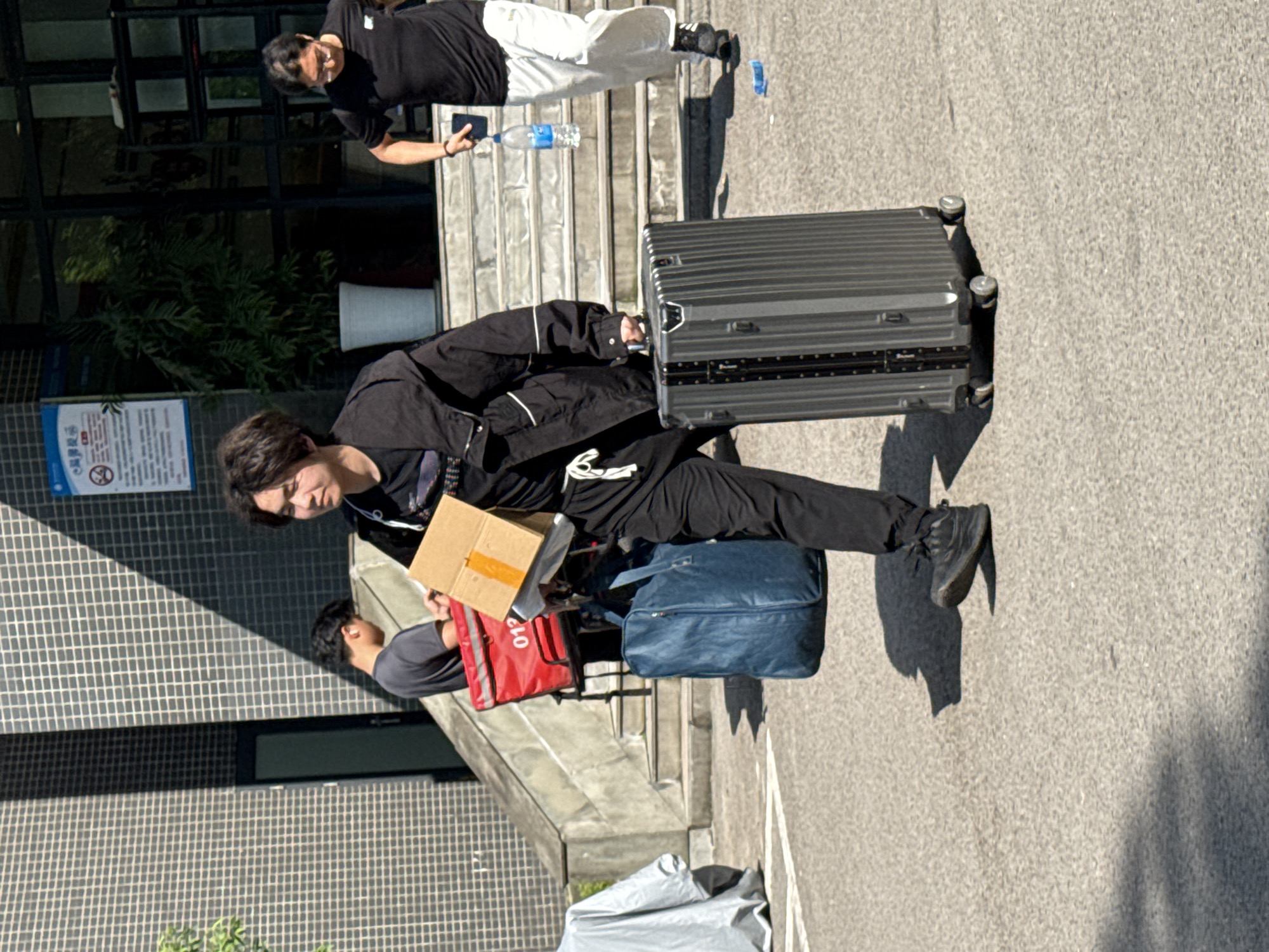 A person is standing on a concrete surface, gripping the handle of a large, metallic, wheeled suitcase to their right. The person is wearing black pants, a black jacket, and black shoes. They are also carrying a small cardboard box and a blue duffel bag. To their left, a person in a gray shirt holds a red food delivery bag. Another person, wearing a black shirt and white pants, stands on the steps behind them. In the background, there is a building with glass windows, a potted plant, and a sign. The scene appears to be outdoors, possibly in Jiangjin, China.