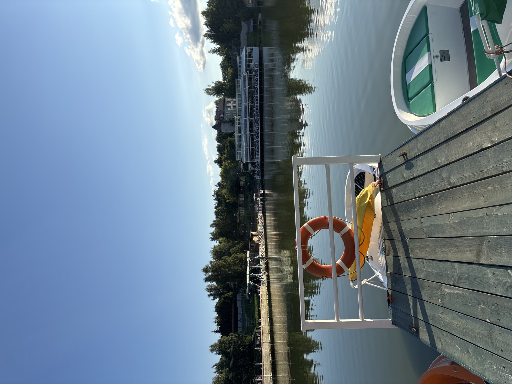 A still, daytime, outdoor shot captures a serene lake scene in Romashkovo, Russia. The foreground features a wooden pier with two white and green pedal boats docked. A bright orange life preserver hangs on a white railing, attached to a smaller yellow float. The calm water reflects the clear blue sky and the lush green trees lining the opposite shore. In the distance, a long, white building with a covered outdoor dining area sits near a large, stately house with a tower. Sunlight casts long shadows across the pier, indicating the late afternoon. The overall atmosphere is peaceful and idyllic.
