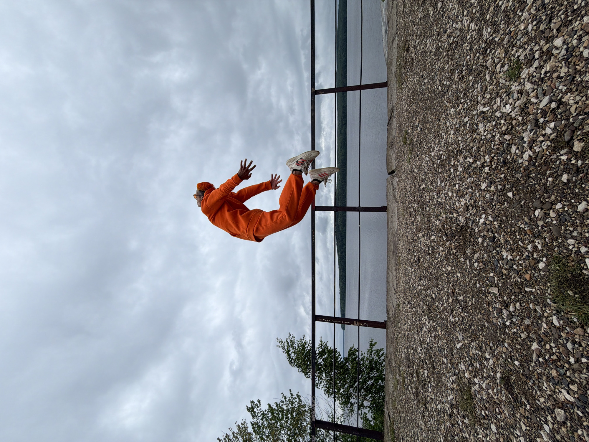 A person in an orange tracksuit and white sneakers is performing a stunt over a railing on a concrete platform. The person is oriented horizontally in the image with their head pointing towards the left and feet towards the right. Behind the railing is a body of water. The sky above is overcast with clouds. Below the railing, trees are visible. The image was taken in the city of Chaikovsky, Russia.