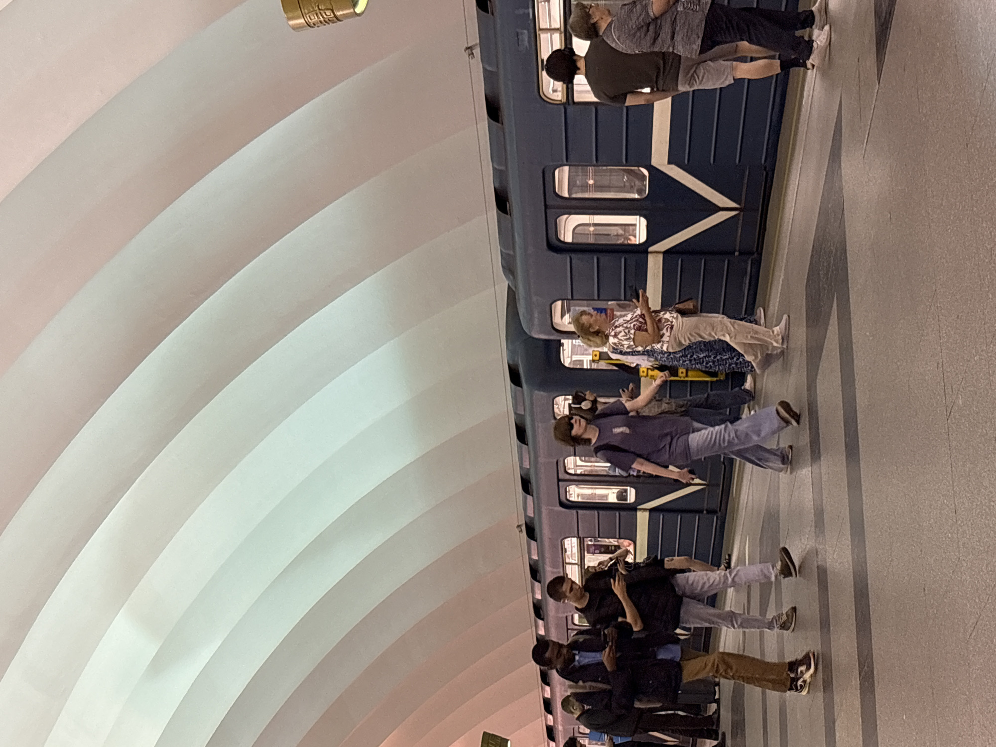 The image captures a scene at a subway station in Saint Petersburg, Russia, likely during daytime. The station features a distinctive arched ceiling with a gradient of pastel colors, ranging from white to pink. A blue subway train is stopped at the platform.
Several people are visible on the platform, some walking and others standing. On the left, a Black man in a dark jacket and blue shirt is looking at his phone. Next to him, a man in a black vest and grey pants is also engrossed in his phone. Further right, a woman in a patterned shirt and light-colored pants is looking towards the train, holding a phone. A woman in a purple t-shirt and jeans walks past, looking ahead. To the far right, a man in a dark t-shirt and shorts stands facing away from the camera, while a woman in a grey patterned shirt and dark pants is positioned behind him.
The platform is made of concrete with dark grey lines indicating the edge. The overall impression is of a busy but orderly metro station, with passengers engaged with their devices while waiting for or disembarking from the train.