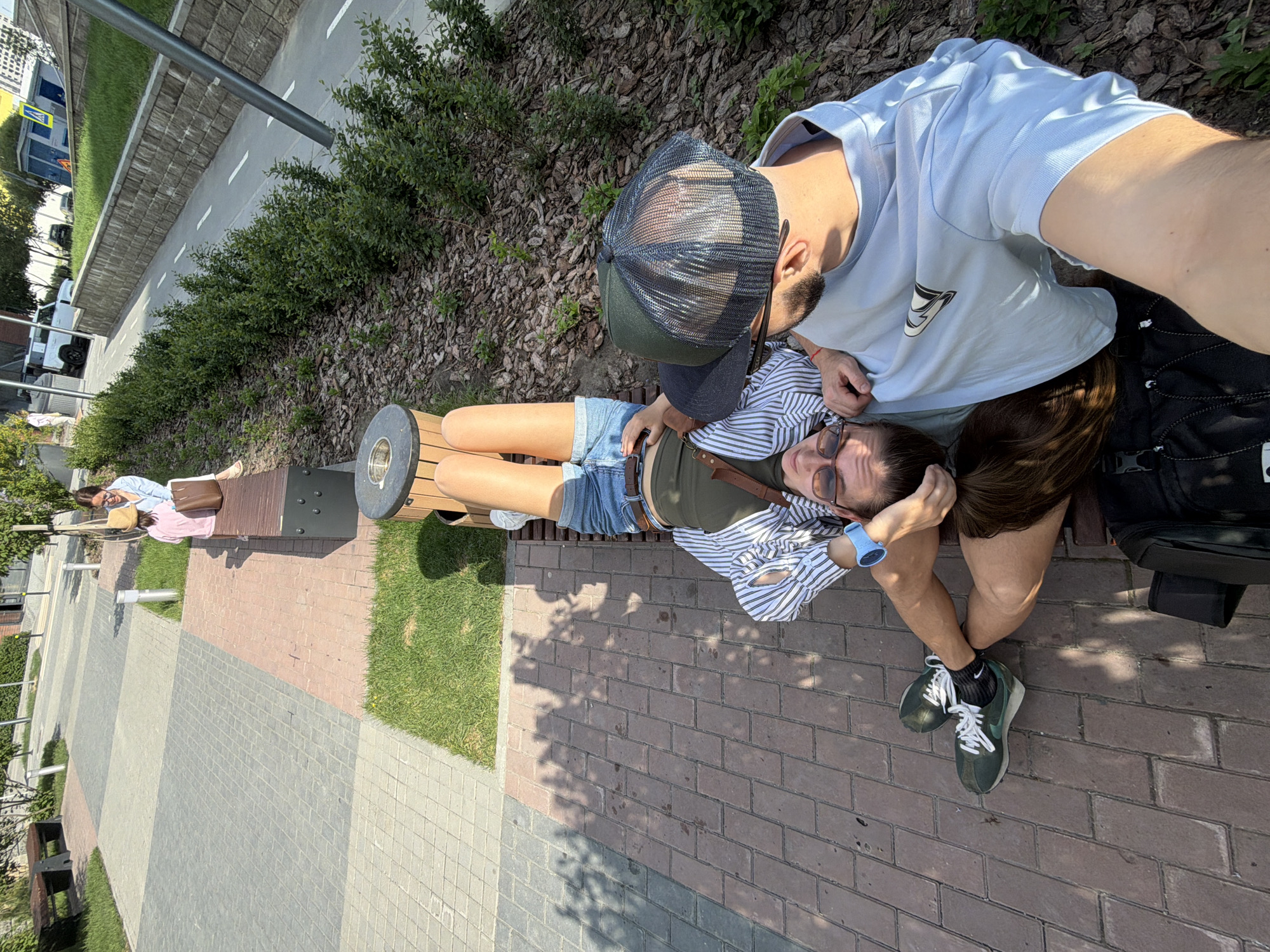 A medium shot from a high angle shows a man and a woman on a modern bench in Tyumen, Russia. The bench is constructed from light brown timber planks and dark gray metal supports. The woman is reclining on the bench with her head resting in the lap of the man, who is seated at the end of the bench. The woman is wearing denim shorts, a striped shirt, and a dark green undershirt with brown leather strap detailing. She also wears dark sunglasses and a light blue smartwatch. The man wears a light blue t-shirt and a dark trucker-style cap. Both appear to be young adults. The ground is covered in red brick pavers, with interspersed sections of gray concrete pavers. A narrow strip of grass borders the pavers, and behind that, a hedge separates the area from a street. In the distance, other pedestrians are seated on similar benches. A street with cars can be seen in the background.