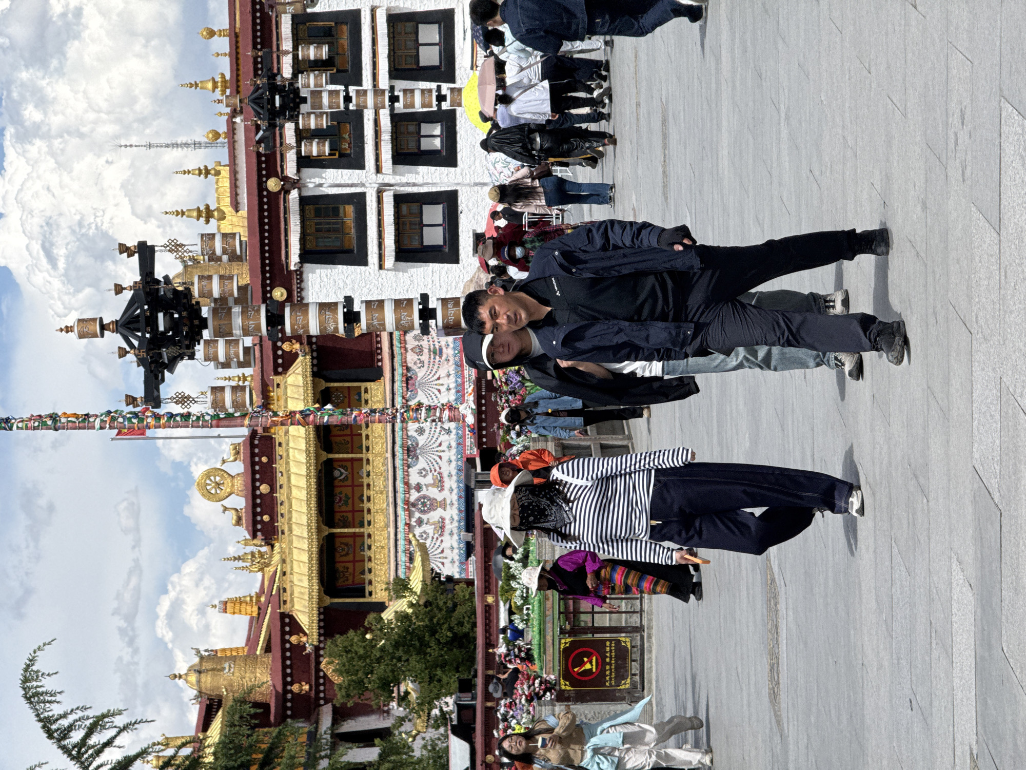The image depicts a street scene in Chengguan District, China, in front of a Tibetan-style building. Several people are visible on the street. One person is wearing a black and white striped shirt, and another pair is wearing black jackets and dark pants. The architecture of the building shows intricate gold details and a white upper level with dark-framed windows. Prayer flags are strung across the scene. The environment includes a paved street, a few trees, and a cloudy sky.