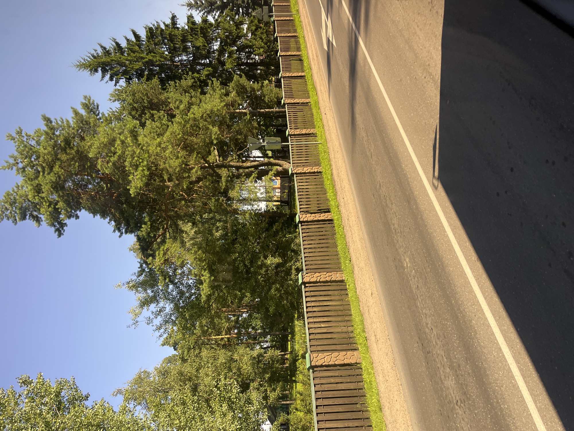 The image depicts a roadside scene in Pastavy, Belarus. A paved road with a white lane divider runs diagonally from the bottom right to the upper left of the frame. To the left of the road is a dirt shoulder and a wooden fence with stone pillars. Beyond the fence, there is lush green grass, trees, and a glimpse of a building. The sky is clear and blue, indicating a sunny day. The overall impression is of a rural or suburban setting in the city of Pastavy.
