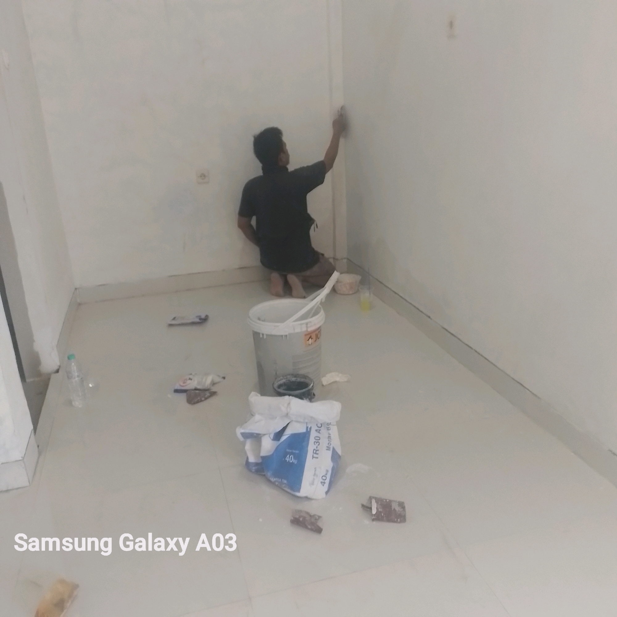 The image depicts an indoor scene, likely a room under renovation or construction. A person, dressed in a dark t-shirt and shorts, is kneeling on the floor and applying a white substance to a wall with a tool, possibly plaster or paint. Various construction materials are scattered around the floor, including a large white bucket containing white sticks, a smaller dark container, and a bag labeled "TR-30 AC 40kg". A plastic water bottle is also visible on the floor. The walls are painted a light color, and there is a baseboard along the bottom of the walls. A power outlet is visible on the wall. The floor appears to be tiled or polished concrete. The text "Samsung Galaxy A03" is overlaid on the bottom left of the image, which is a likely watermark from the device used to capture the photograph, suggesting the location might be a residential or commercial space where this phone is used. The image does not provide any specific visual cues to pinpoint the location to Kenanga, Indonesia, other than the provided context.