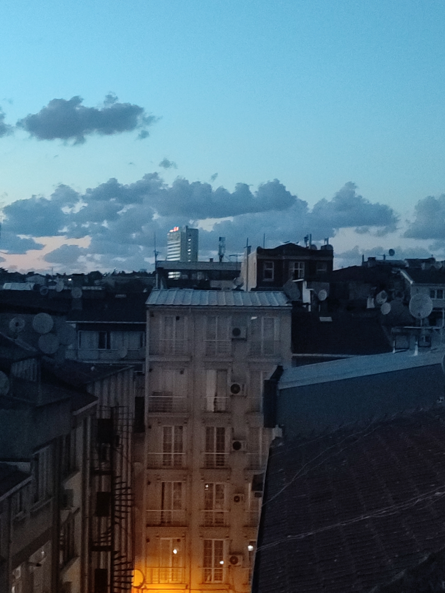 This is a medium-shot, eye-level, vertical, slightly blurry photograph taken at dusk in Kadıköy, Turkey. The sky is a deep blue with scattered gray clouds. The foreground is dominated by the rooftops and upper floors of several apartment buildings. A tall, modern building with illuminated windows and what appears to be a red sign or logo on top is visible in the distance, to the left of center. Several satellite dishes are visible on the rooftops, as well as exterior air conditioning units on the side of one building. A dark, angled roof occupies the lower right quadrant of the image. A spiral staircase is visible on the side of a building on the left. Warm yellow light emanates from the ground floor of a building on the bottom right. The overall scene depicts an urban environment at twilight.