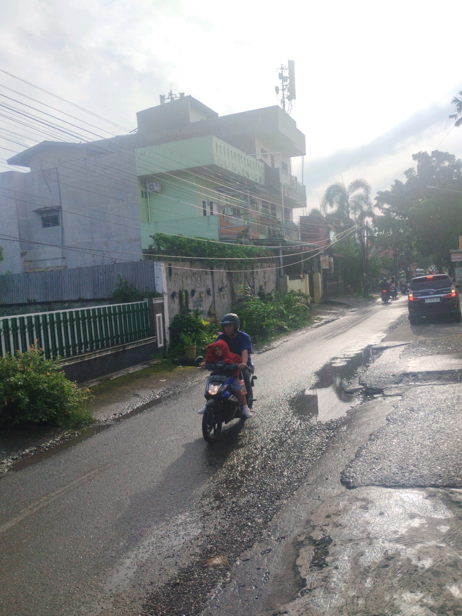 Two people are riding on a motorcycle on a wet asphalt road. The rider, a male wearing a blue shirt and helmet, is driving. A female, wearing a red hooded jacket and possibly a helmet underneath, is sitting behind him. The motorcycle has a license plate that reads "BB 8015 YG". The road is in disrepair, with visible potholes and puddles. On the left side of the road, there is a green metal fence in front of a building. The building appears to be a multi-story structure with a light green facade. Power lines crisscross overhead. On the right side of the road, a dark-colored SUV is parked. In the background, trees and more buildings are visible, as well as a cell tower on top of one of the buildings. The sky is overcast with bright patches, suggesting it is daytime. The scene depicts a typical street in the City of Medan, Indonesia, characterized by its urban environment, residential buildings, and infrastructure challenges like poorly maintained roads.