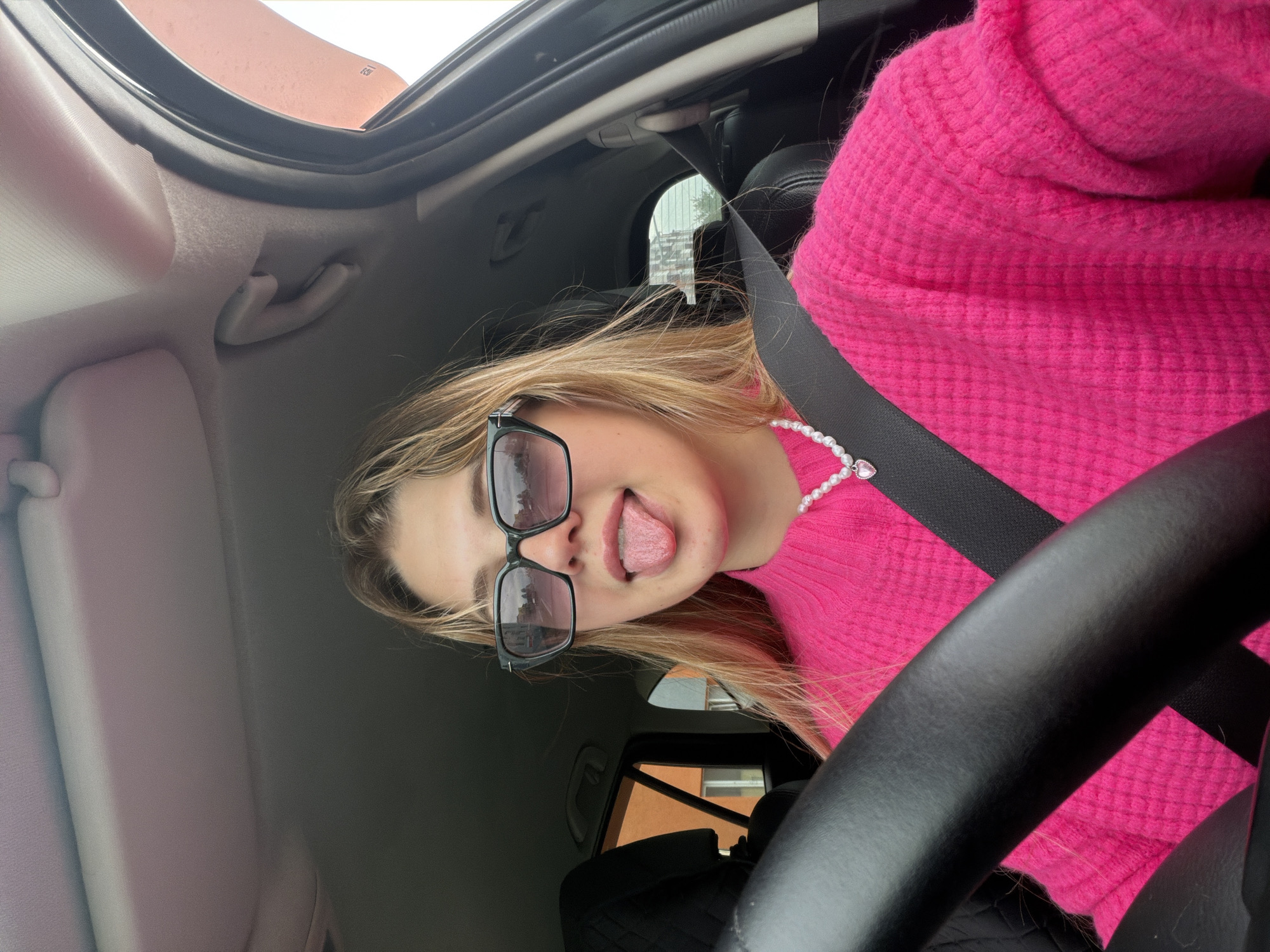 A young woman is sitting in the driver's seat of a car, wearing a bright pink sweater, black sunglasses, and a pearl necklace with a heart pendant. She is sticking her tongue out. She is wearing a seatbelt. The interior of the car is visible, including the steering wheel and dashboard. The car appears to be parked. Based on the objects in the background, this image was not taken in Agryz, Russia.